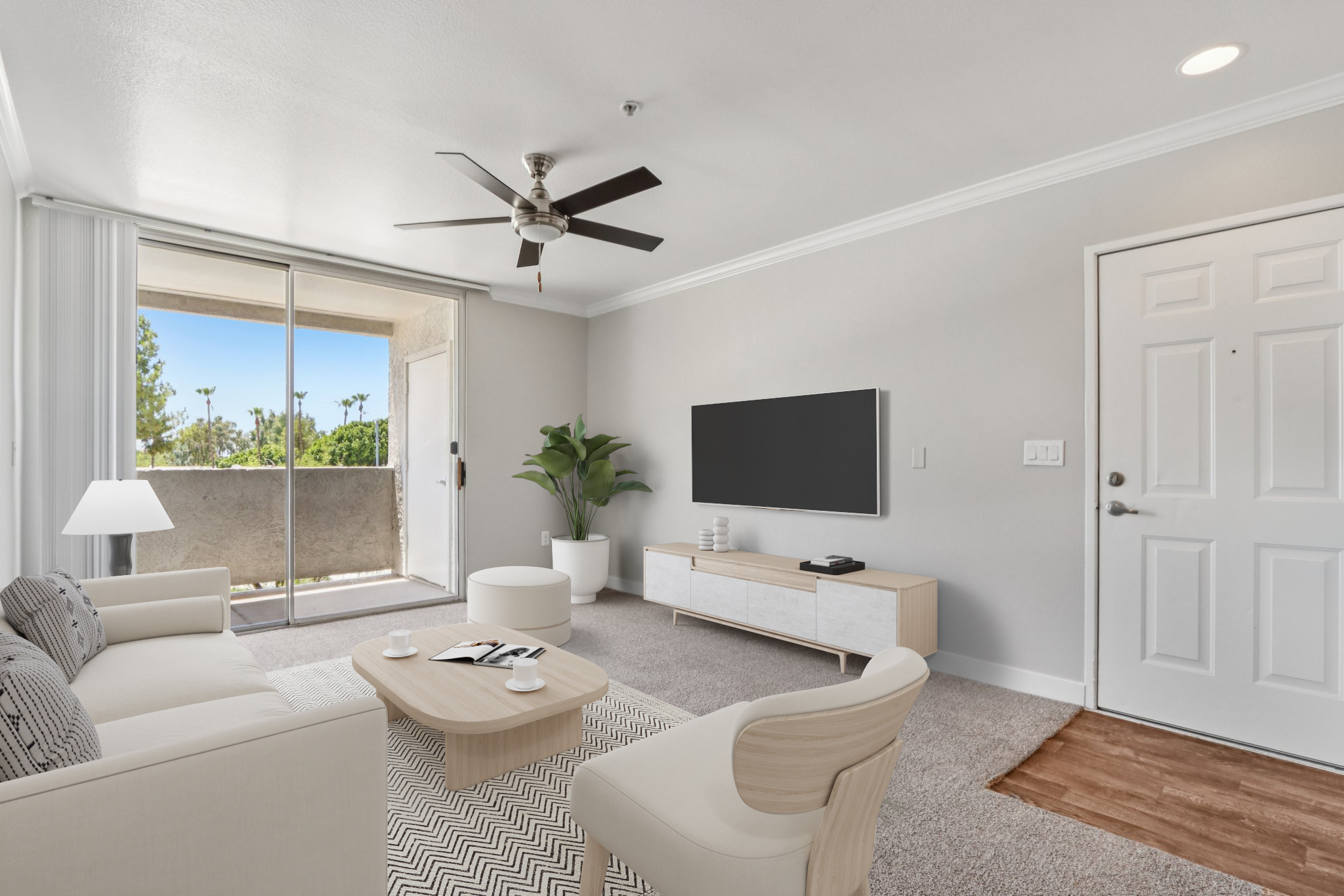 A modern living room featuring a light-colored sofa, a coffee table, and a stylish entertainment unit with a flat-screen TV. A ceiling fan hangs above, and a sliding glass door leads to a balcony with outdoor greenery. The space is bright and airy with neutral tones and a cozy ambiance.