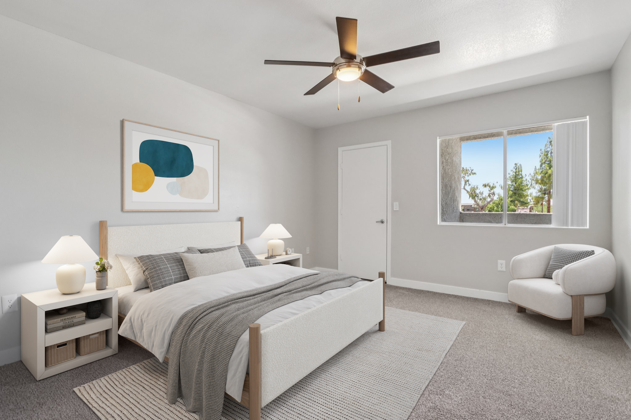 A modern bedroom featuring a light-colored bed with gray and white bedding, a nightstand with lamps on each side, and a cozy reading area with a small armchair. The walls are painted in a soft gray, and there's a large window allowing natural light to illuminate the space. A piece of abstract art decorates the wall above the bed.