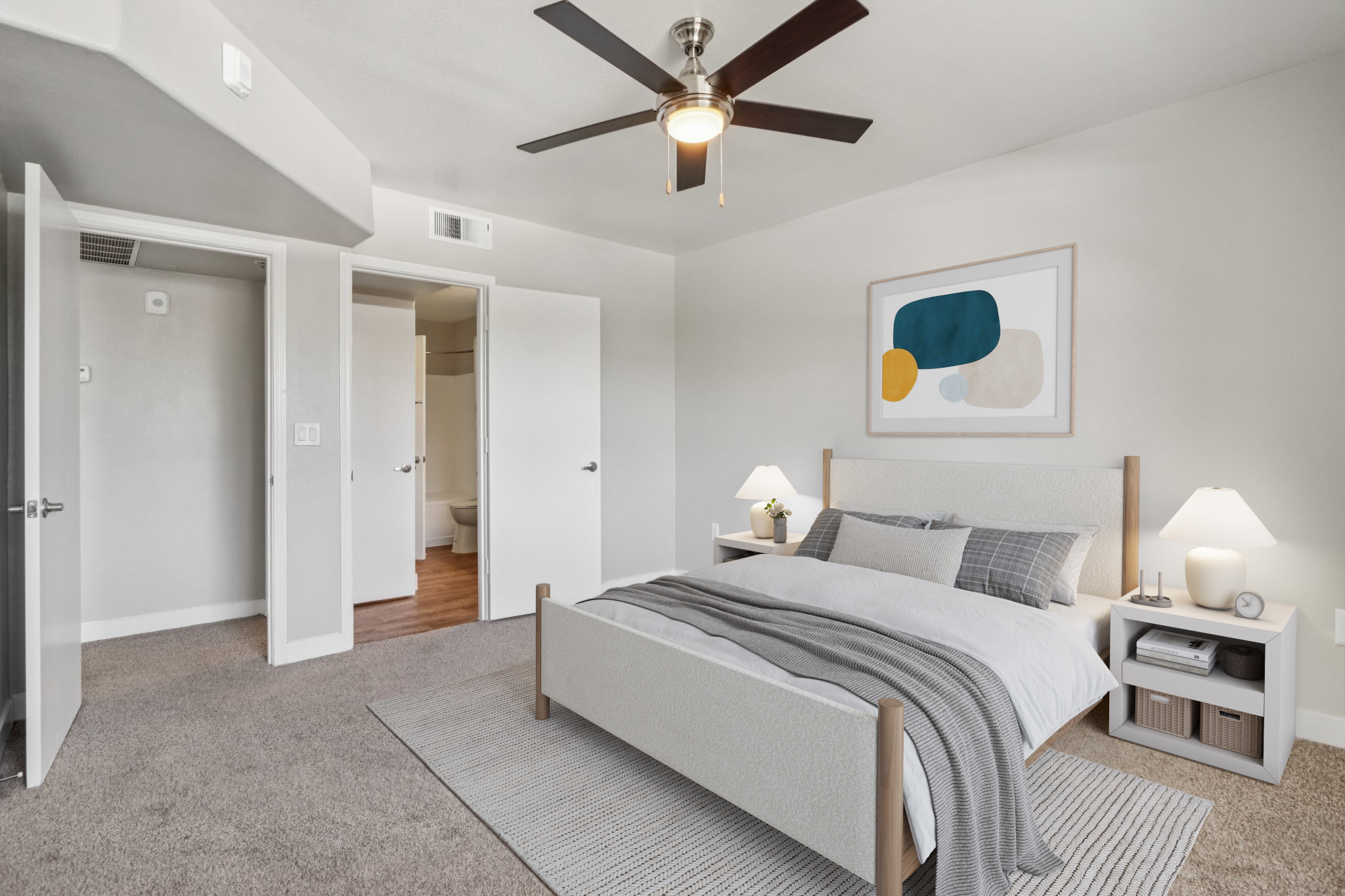 A modern bedroom featuring a queen-sized bed with white and gray bedding, two bedside tables with lamps, and a large area rug. There is a ceiling fan, a piece of abstract wall art, and a doorway leading to a bathroom. The walls are light-colored, and the space is well-lit, creating a cozy atmosphere.