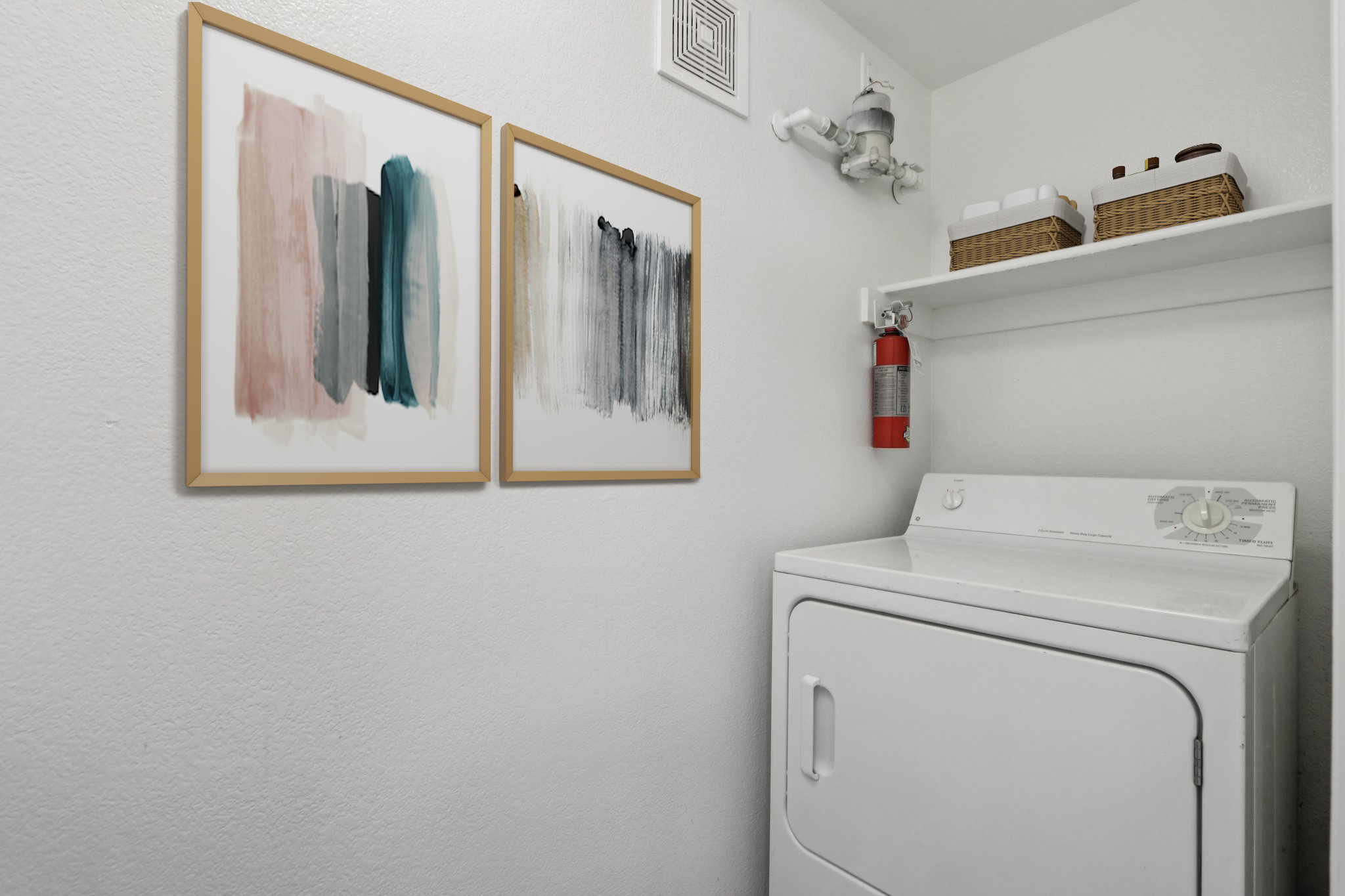 A laundry room featuring a white washing machine and two framed abstract artworks on the wall. The room has a clean, minimalist design with a shelf displaying decorative baskets and a fire extinguisher mounted nearby. The walls are painted in a neutral color.