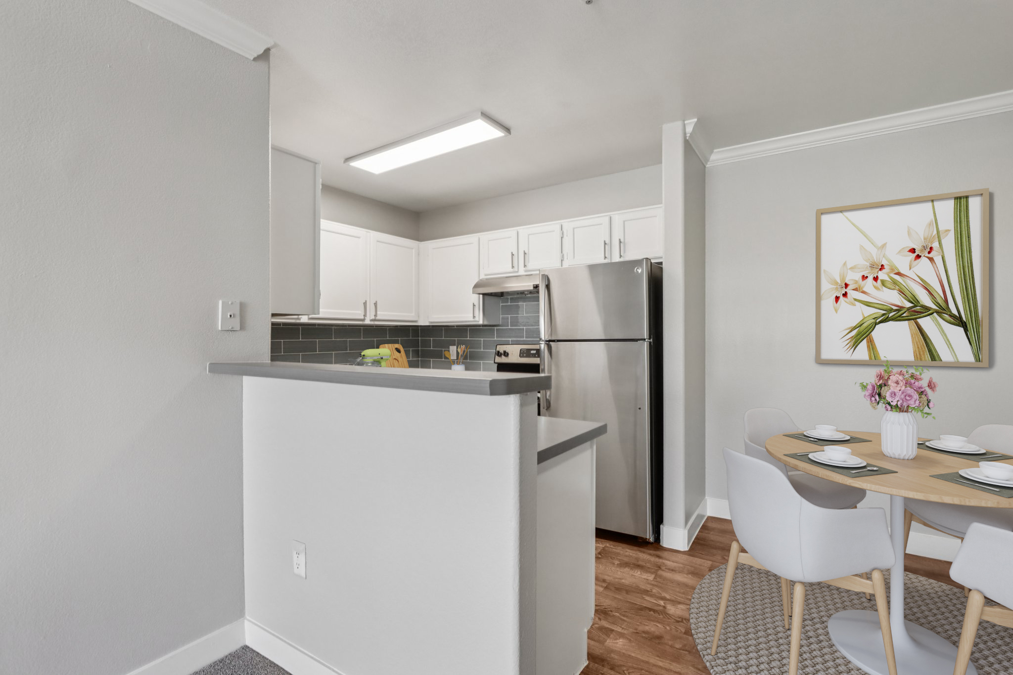 A modern kitchen with white cabinets and stainless steel appliances. The space features a gray countertop and a dining area with a round table set for four, accompanied by white chairs. A framed floral artwork is on the wall, and the flooring is a warm wood tone. Natural light illuminates the area.