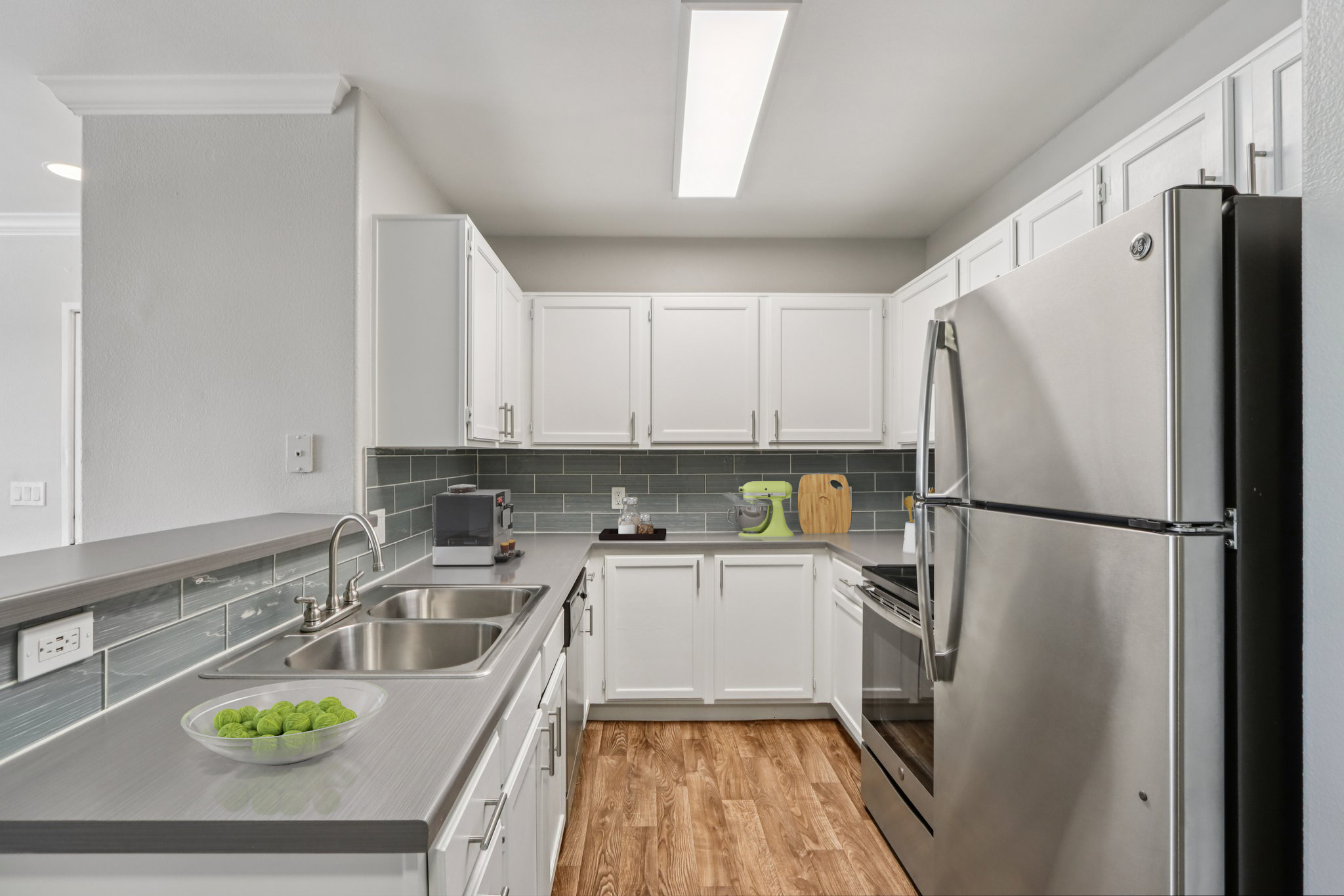 A modern kitchen featuring white cabinets, a stainless steel refrigerator, a gray countertop, and a light-colored wood floor. The backsplash is a subtle gray tile, and a bowl of green apples sits on the counter. The space is well-lit with overhead lighting, creating a clean and inviting atmosphere.