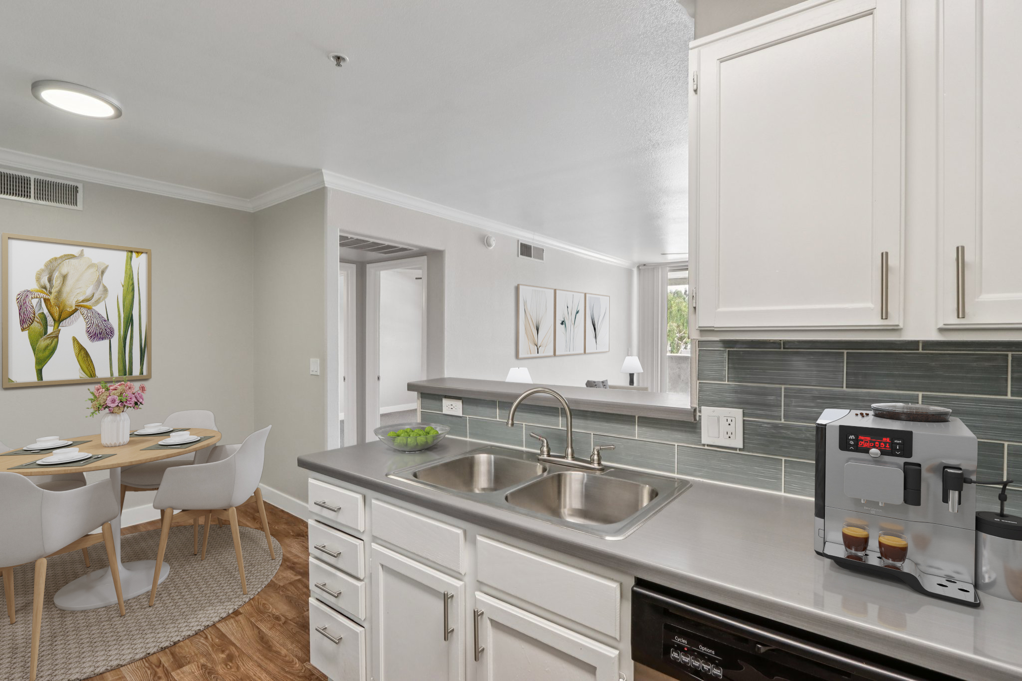 A modern kitchen featuring white cabinetry, a stainless steel sink, and a coffee machine. There is a small dining area with a round table and light-colored chairs, complemented by a decorative floral artwork on the wall. Natural light enters through nearby windows, enhancing the inviting atmosphere.