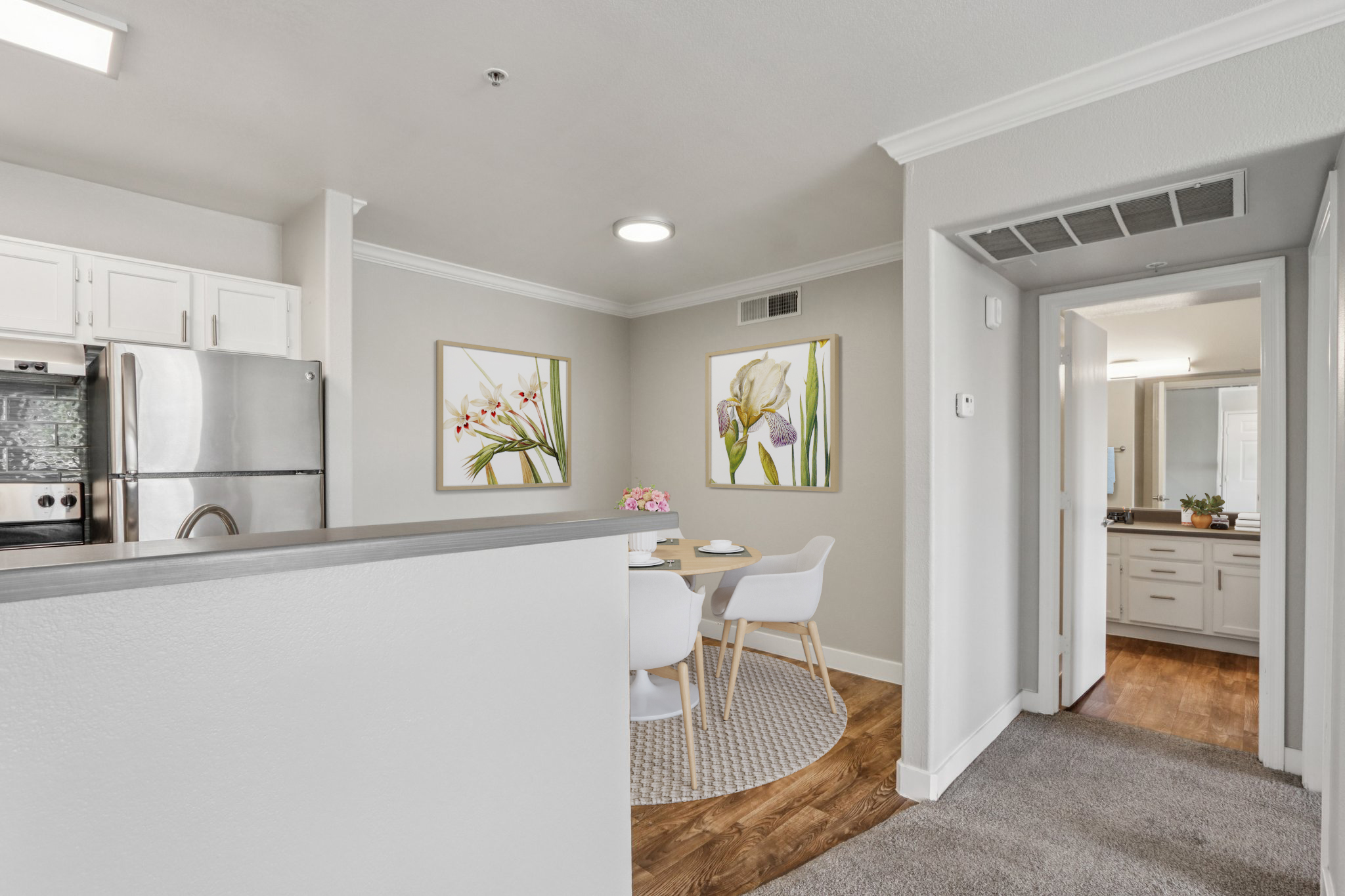 A modern kitchen and dining area featuring a stainless steel refrigerator, white cabinetry, and a round dining table with two chairs. The walls are decorated with floral artwork, and there is a glimpse of a bathroom in the background, providing a bright and inviting space.