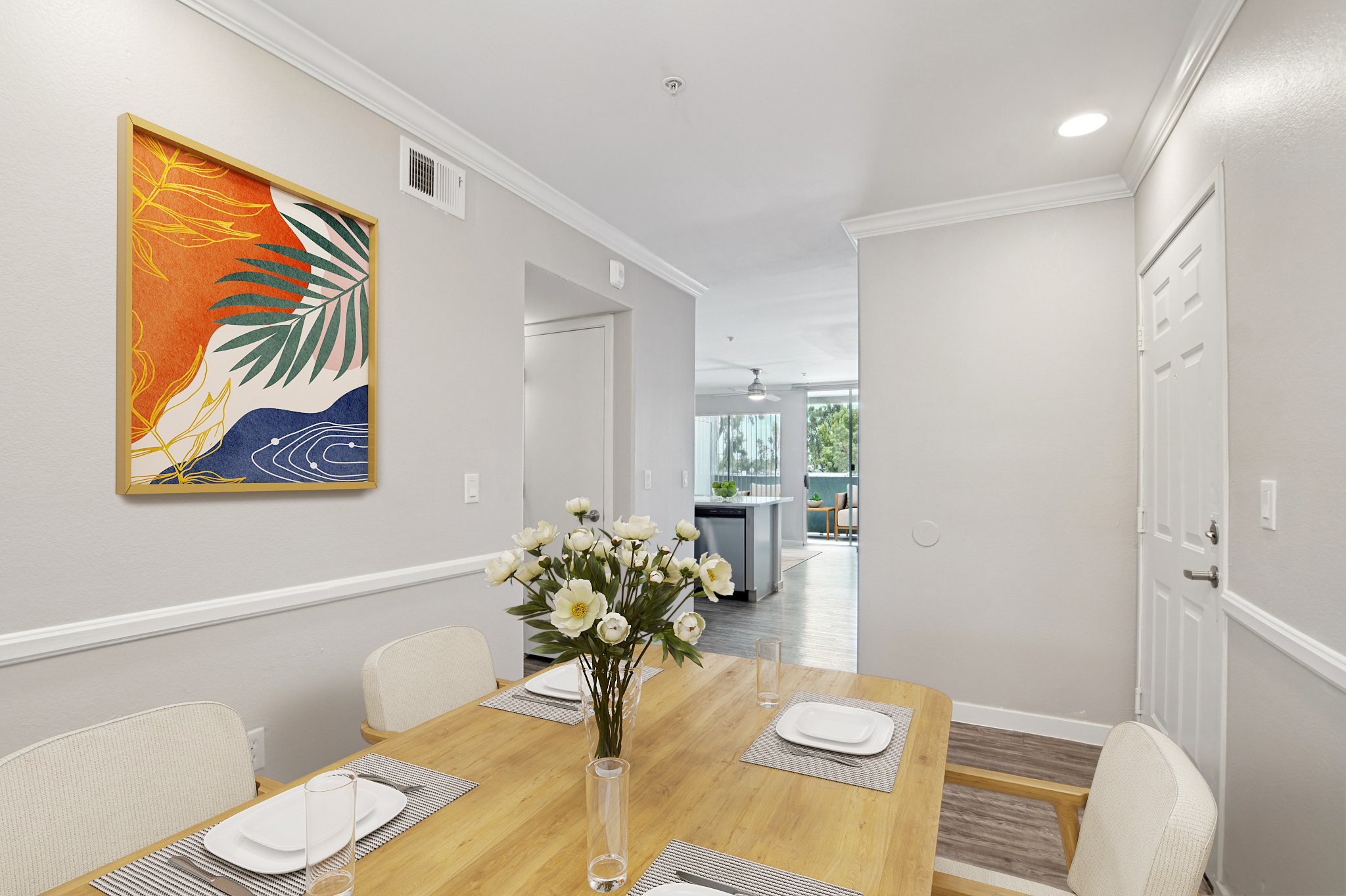 A modern dining area featuring a wooden table set with white dishes and glasses, accompanied by a vase of white flowers. A vibrant abstract artwork with orange, green, and blue elements is displayed on the wall. Bright, neutral walls and a doorway lead to a light-filled living space in the background.