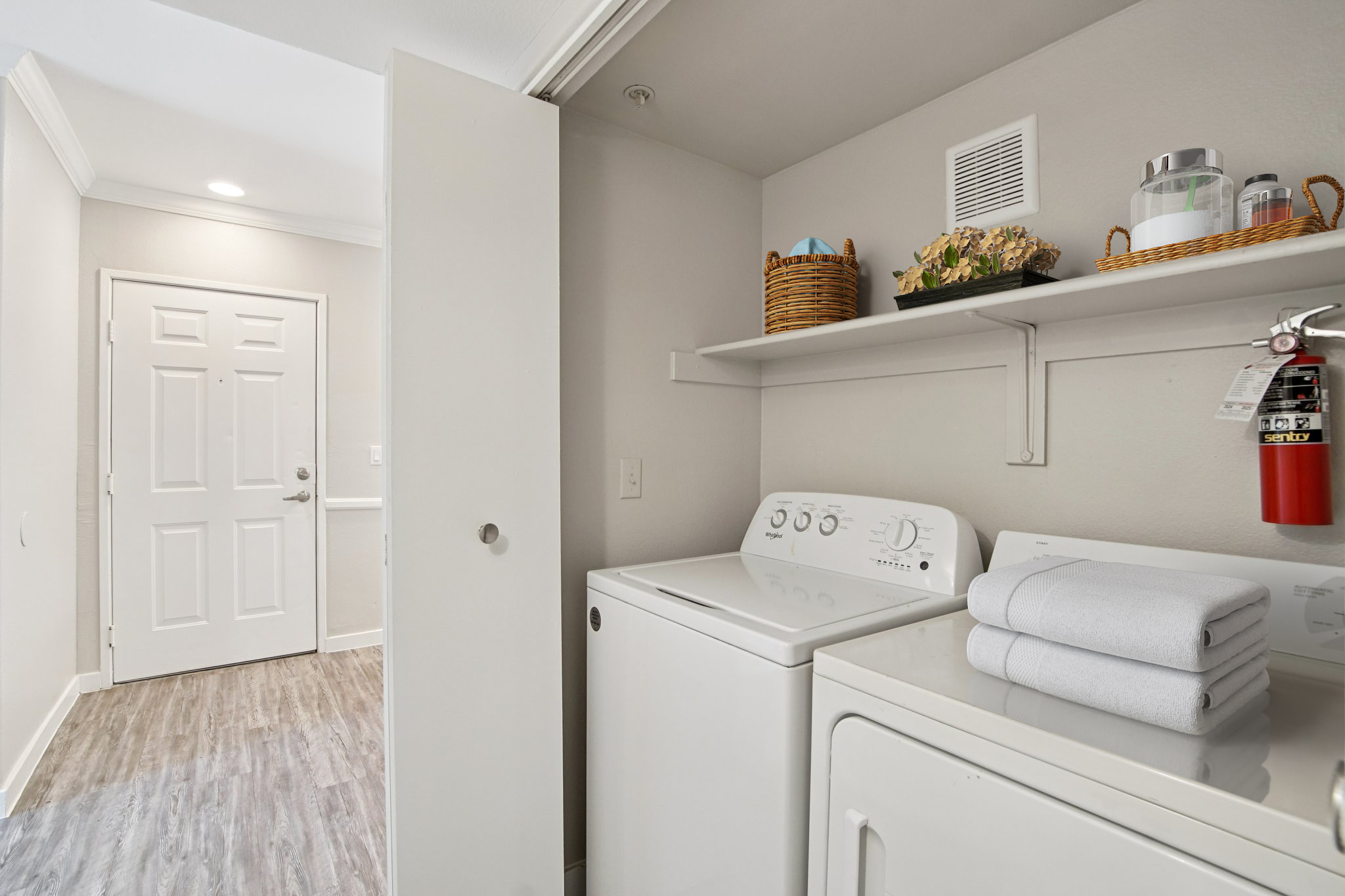 A clean laundry room featuring a white washing machine and dryer, neatly placed towels, and shelves with storage baskets. A door is visible in the background, leading to another room, and a fire extinguisher is mounted on the wall for safety. The walls are painted in neutral tones, enhancing the bright atmosphere.