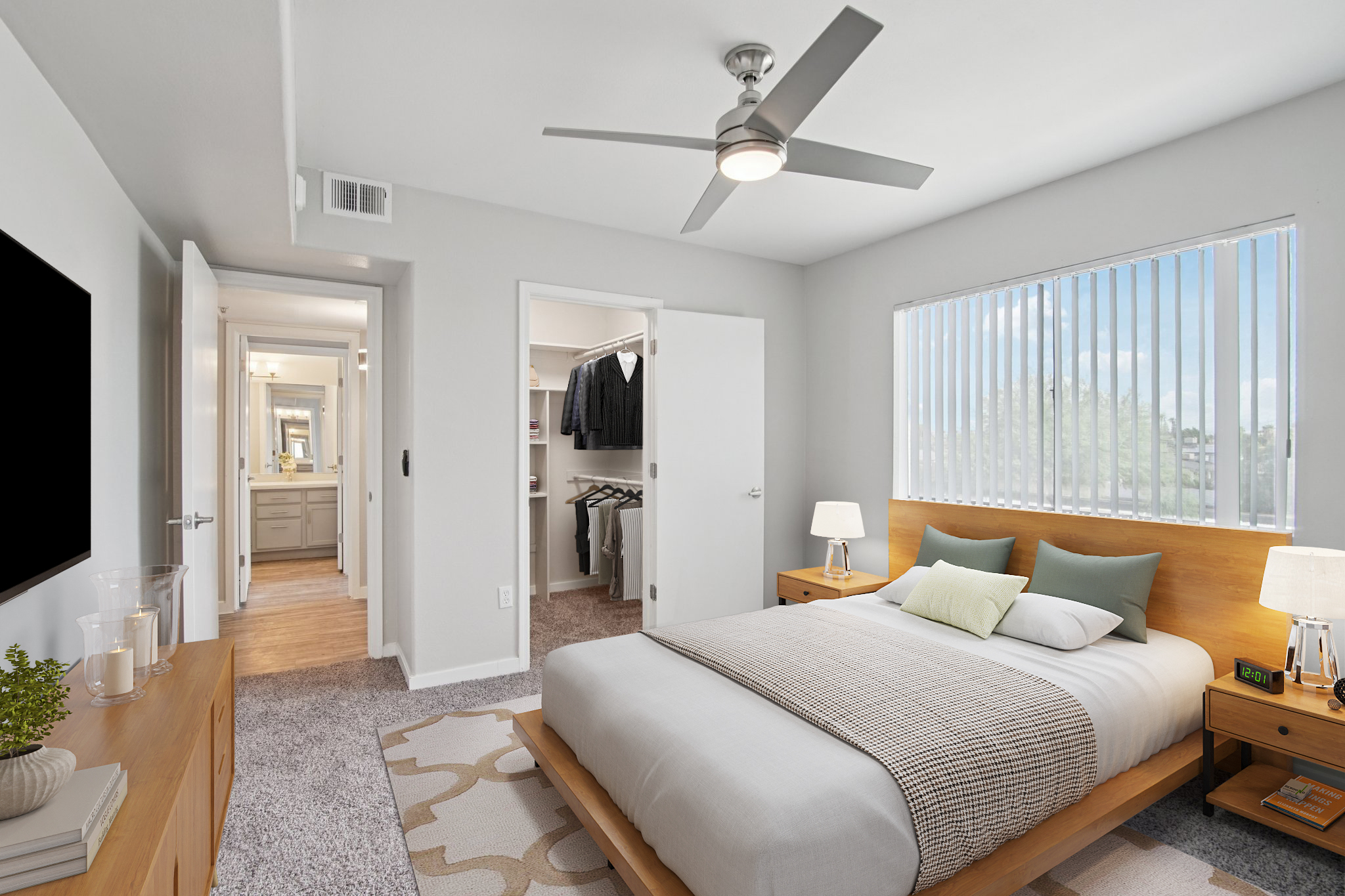 Bright and modern bedroom featuring a queen-sized bed with a gray and beige bedding setup. There are wooden nightstands on either side with lamps. A ceiling fan is overhead, and natural light streams in through a window with vertical blinds. A closet with hanging clothes is visible through an open door.