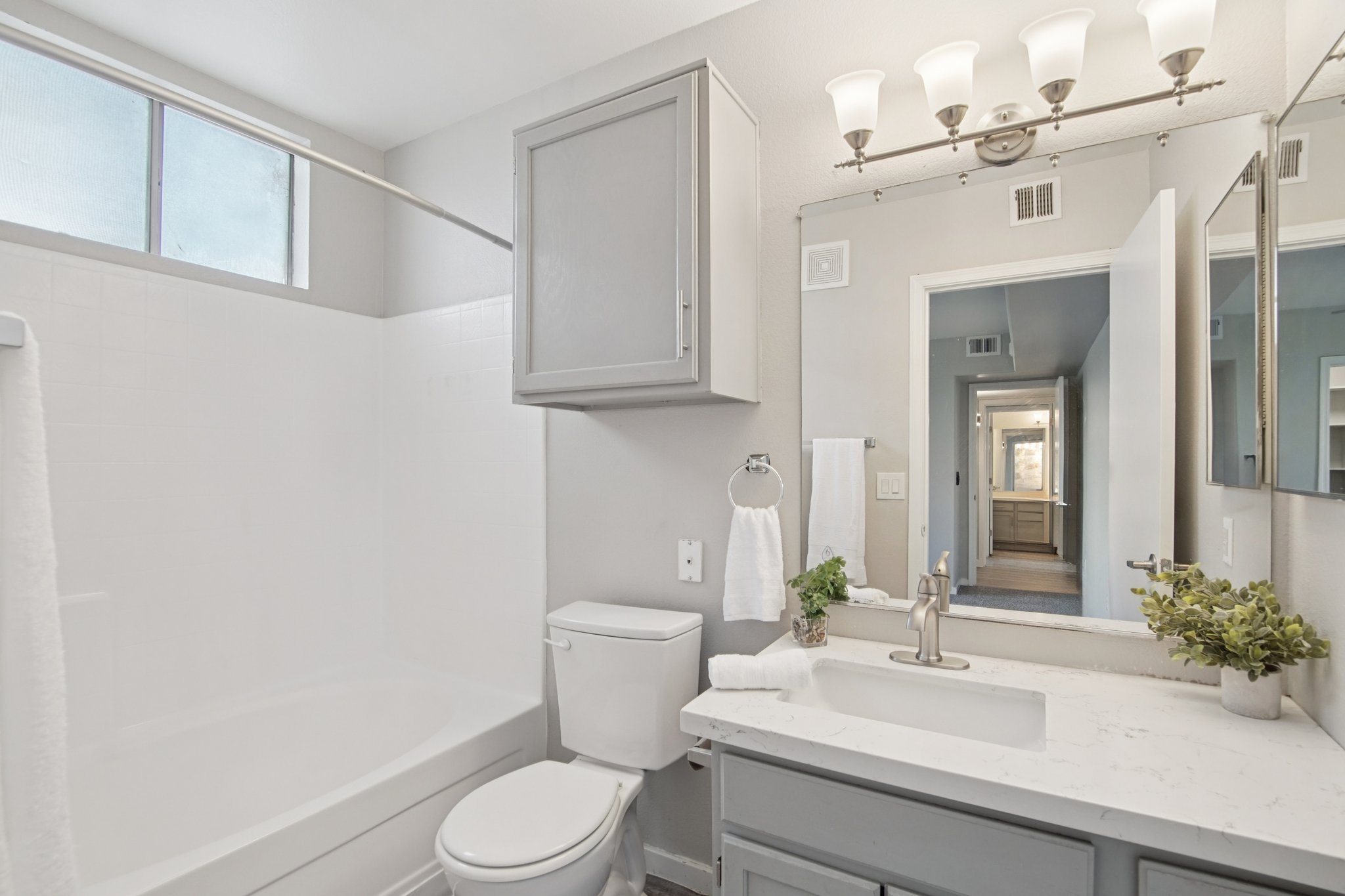 A modern bathroom featuring a white bathtub, a toilet, and a vanity with a marble countertop. Above the vanity, there are four light fixtures. The walls are painted light gray, and there's a small plant on the counter. In the background, a mirror reflects a hallway with carpeted flooring and additional light.
