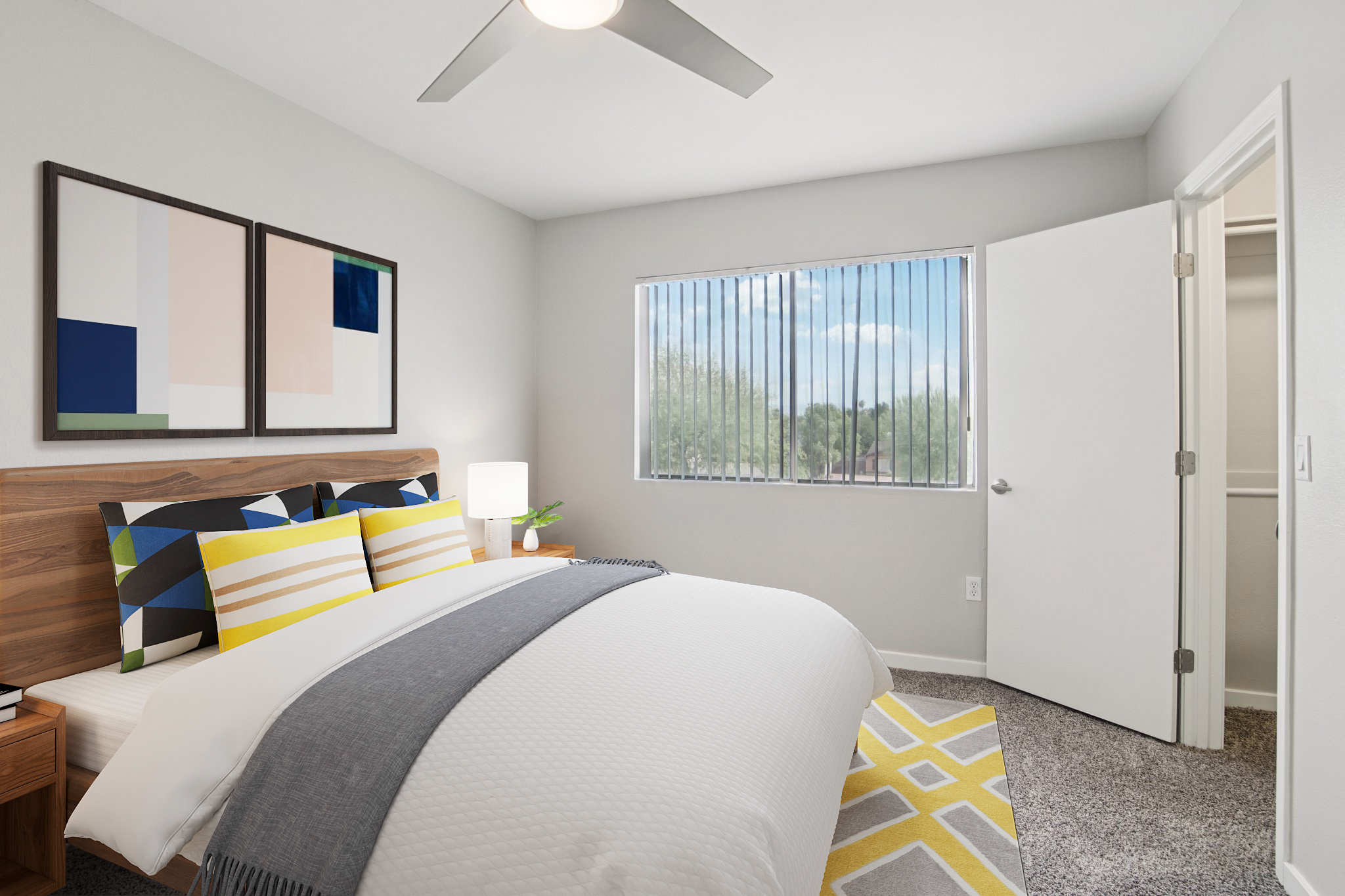A cozy bedroom featuring a queen-size bed with decorative pillows, a modern nightstand with a lamp, and framed artwork on the wall. A large window offers natural light and a view of the outdoors, while a light gray carpet and a geometric area rug complete the inviting atmosphere. An open door leads to a closet space.