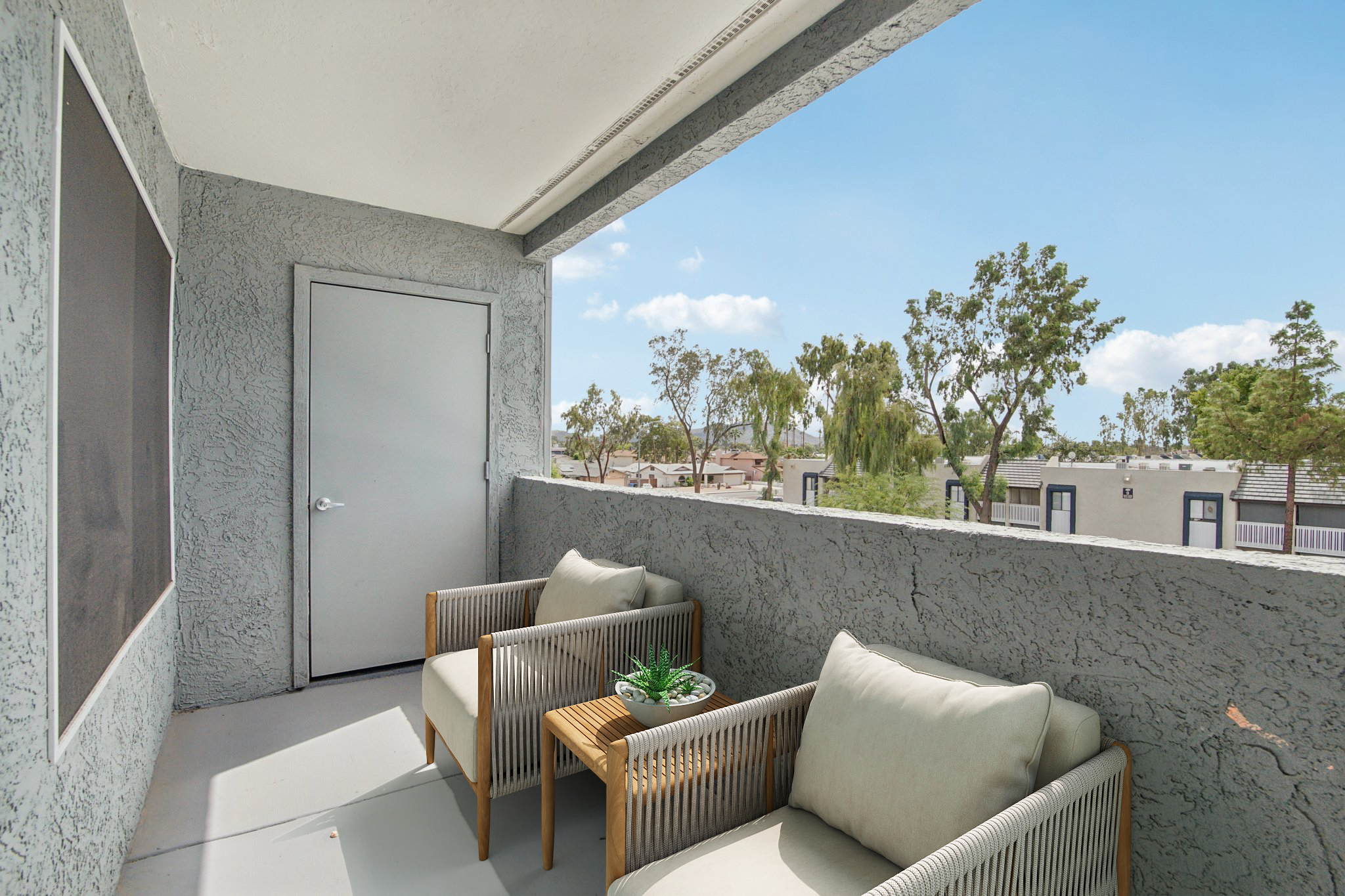 A balcony featuring two comfortable chairs and a small wooden table with a decorative plant. The view includes trees and buildings under a clear blue sky. The walls are textured and light gray, adding to the serene atmosphere.