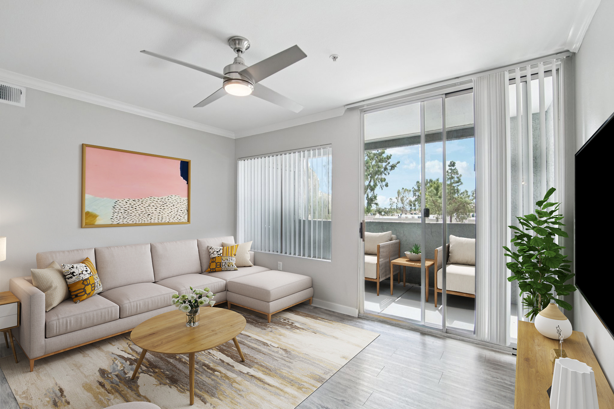 A stylish living room featuring a light-colored sofa with decorative pillows, a round wooden coffee table, and a rug. Large windows with blinds allow natural light, while a ceiling fan adds a modern touch. In the background, there is a balcony with two outdoor chairs and potted plants.