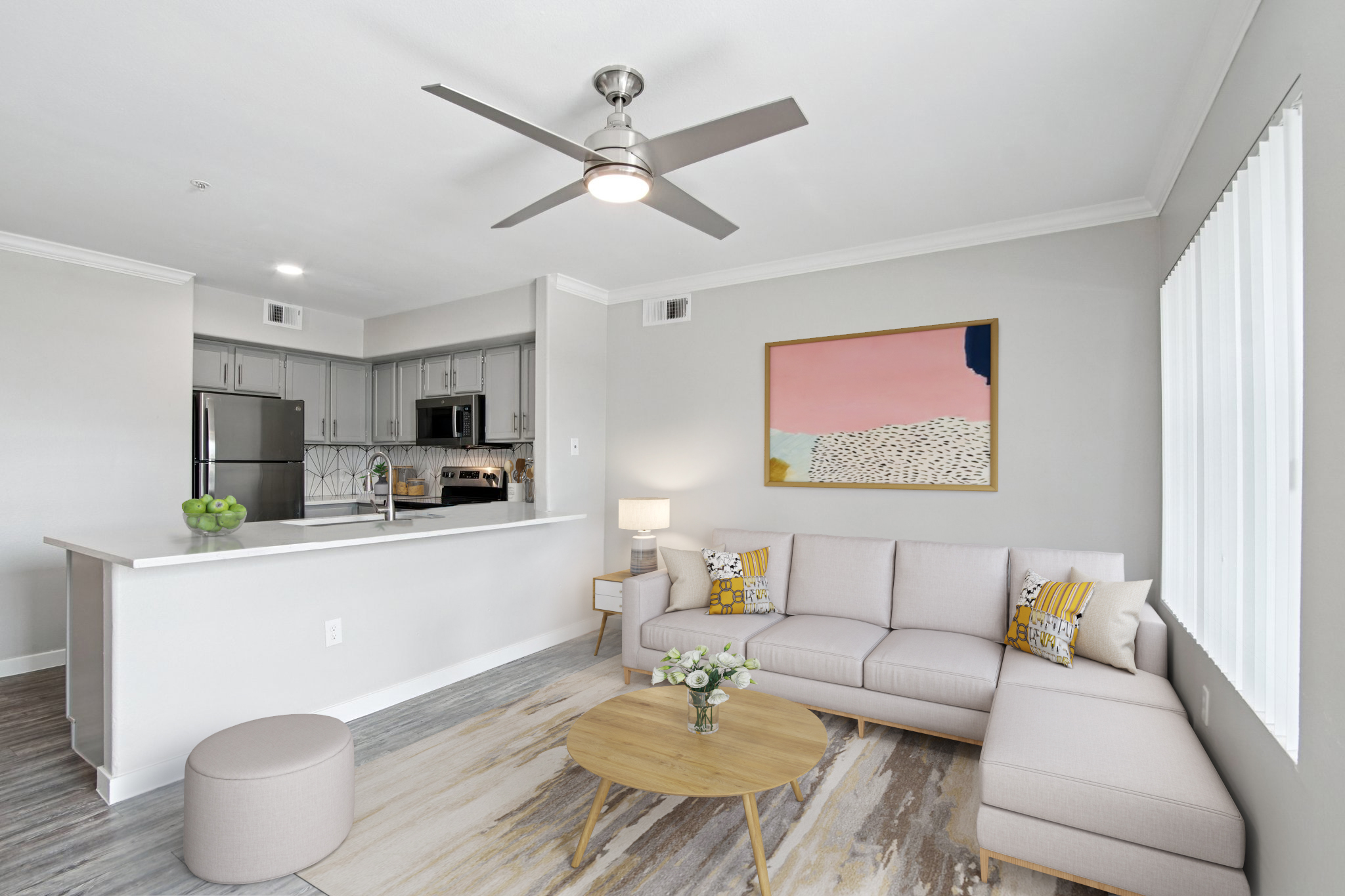 A stylish living room featuring a light-colored sectional sofa with decorative pillows, a round wooden coffee table, and a floor lamp. A kitchen area with modern cabinetry is visible in the background. The walls are painted in soft gray tones, and a colorful abstract artwork hangs on the wall, adding a pop of color.