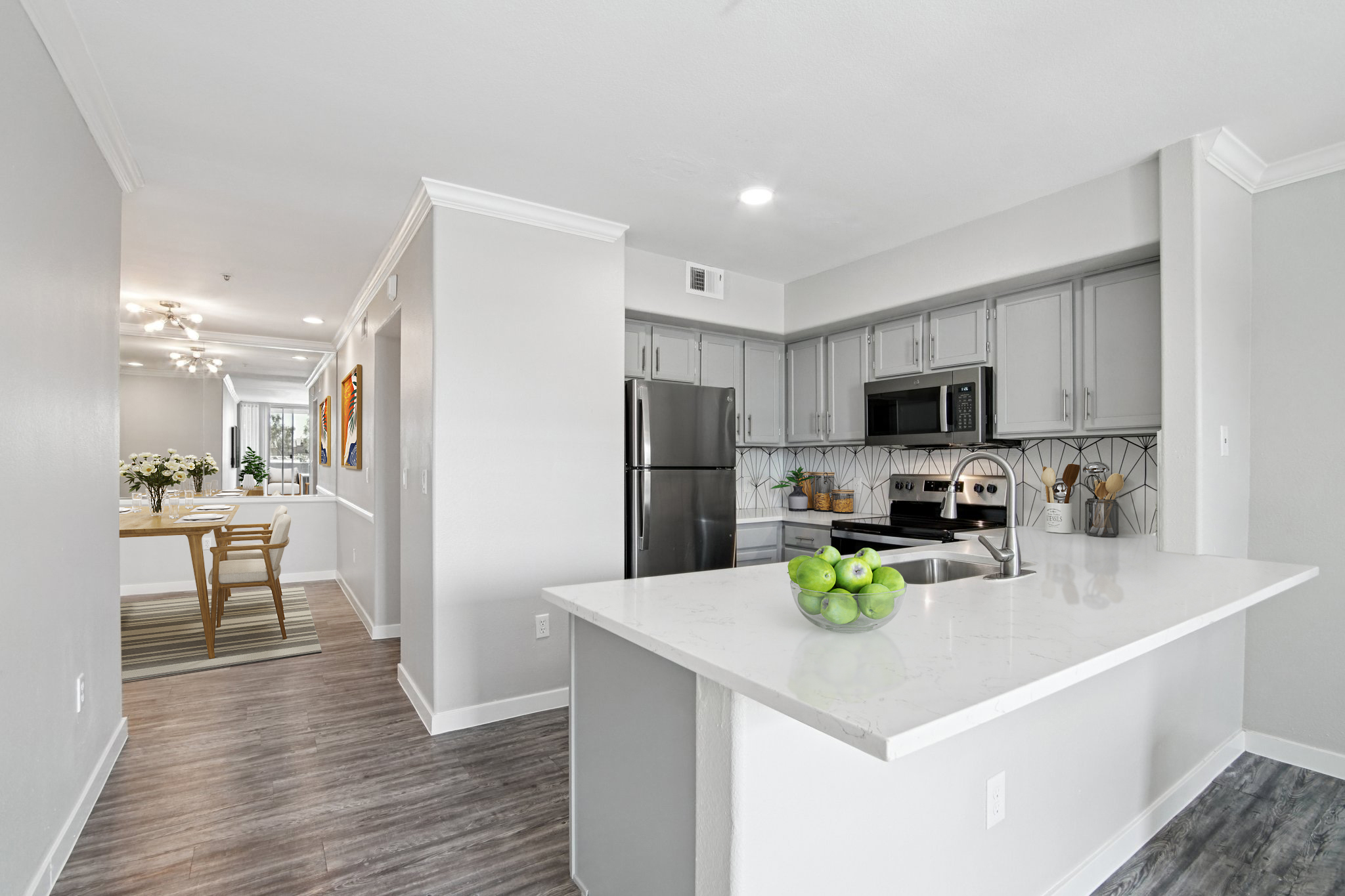 Modern kitchen featuring gray cabinets, stainless steel appliances, and a white countertop. A bowl of green apples sits on the counter. The kitchen is open to a dining area with a wooden table and chairs visible in the background, with light-colored flooring and bright lighting.