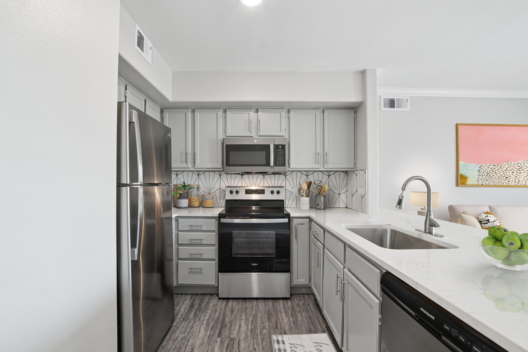 Modern kitchen featuring stainless steel appliances, including a refrigerator, microwave, and oven. The light gray cabinetry complements the stylish backsplash. A sink with a contemporary faucet is visible, along with a countertop adorned with green apples and kitchen utensils. Bright, inviting decor completes the space.