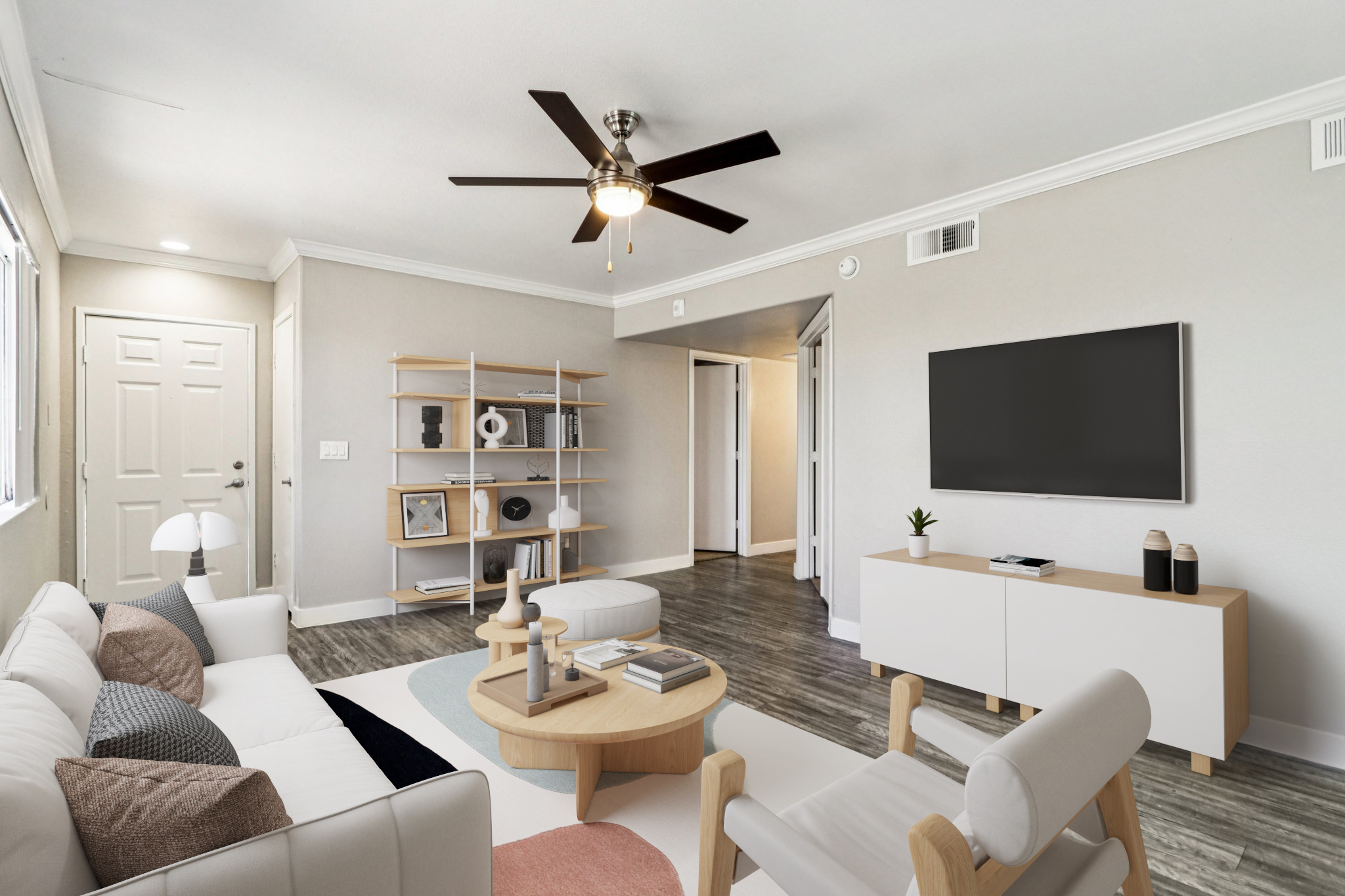 A modern living room featuring a white sofa with colorful cushions, a round coffee table, and a minimalist decor. There’s a wall-mounted TV and an open shelving unit displaying decorative items. A ceiling fan hangs above, and natural light streams in through the windows, creating a bright and inviting atmosphere.