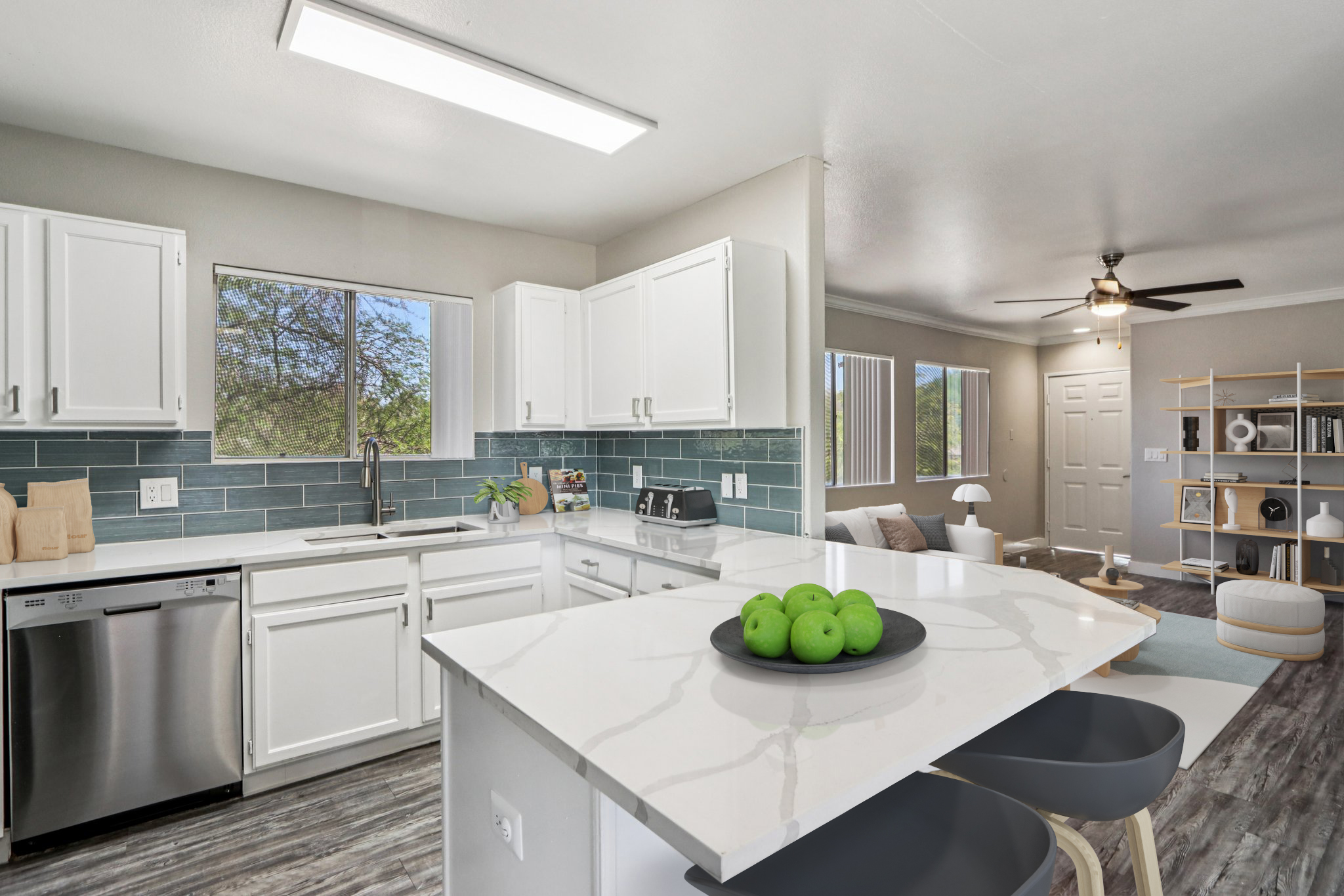 A modern kitchen featuring white cabinets, a marble-patterned countertop, and a stainless steel dishwasher. Green apples in a bowl sit on the island. The space is bright and airy, with large windows, blue-green backsplash tiles, and an adjoining living area visible in the background.
