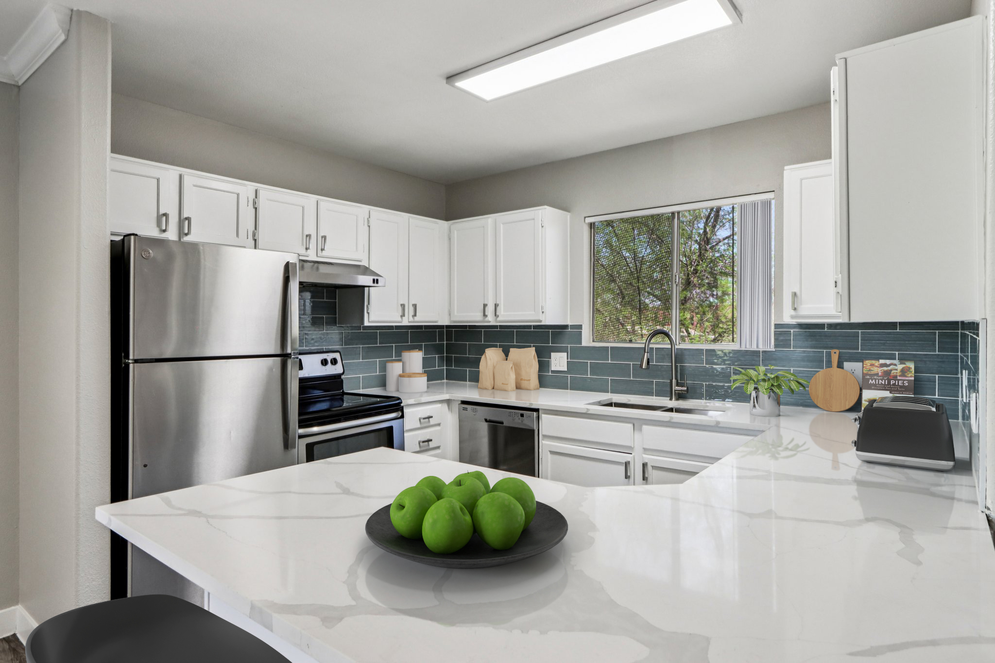 Modern kitchen with white cabinetry, stainless steel appliances, and a large marble countertop. A bowl of green apples sits in the center, and a window offers a view of greenery outside. The backsplash features blue tiles, enhancing the contemporary feel of the space.