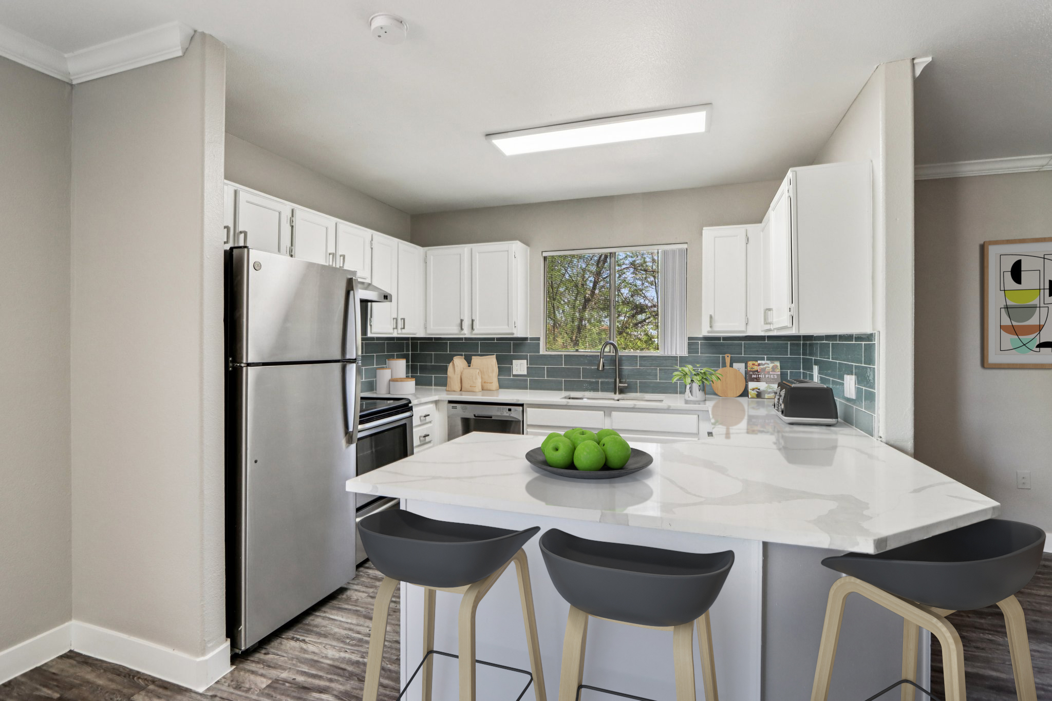 Modern kitchen with stainless steel appliances and a large island featuring a marble countertop. Three black bar stools are positioned around the island. White cabinets and a blue tile backsplash complement the design. A window provides natural light, and a bowl of green apples is on the island.