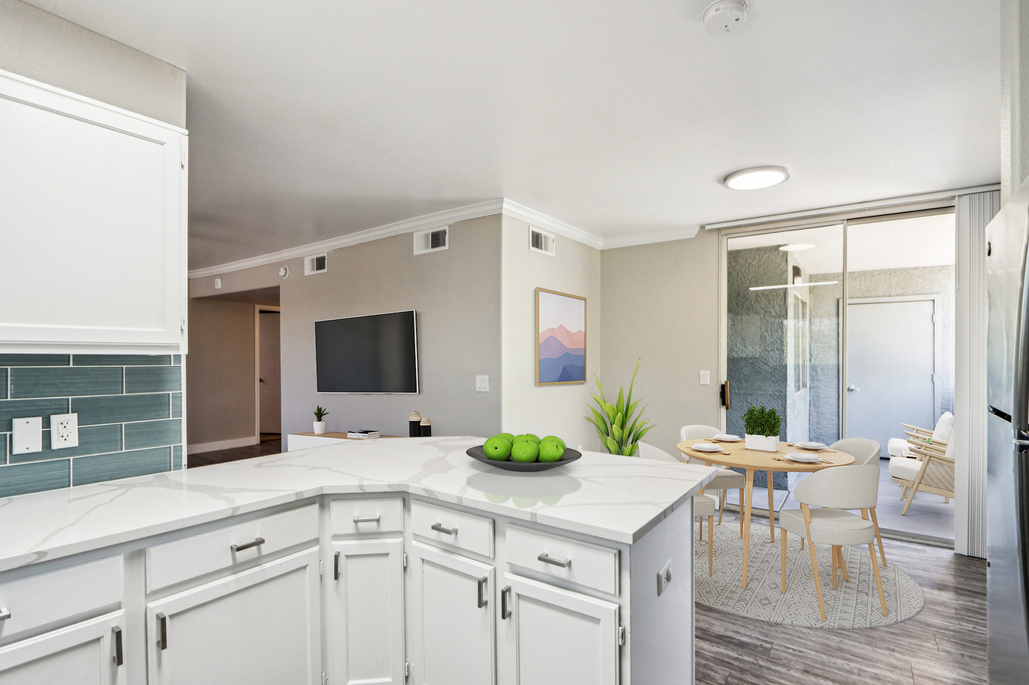 Modern kitchen with white cabinetry and a marble countertop, featuring a bowl of green apples. In the background, a dining area with a round table and light-colored chairs is visible, alongside a living space with a flat-screen TV on the wall. Natural light enters through large windows.
