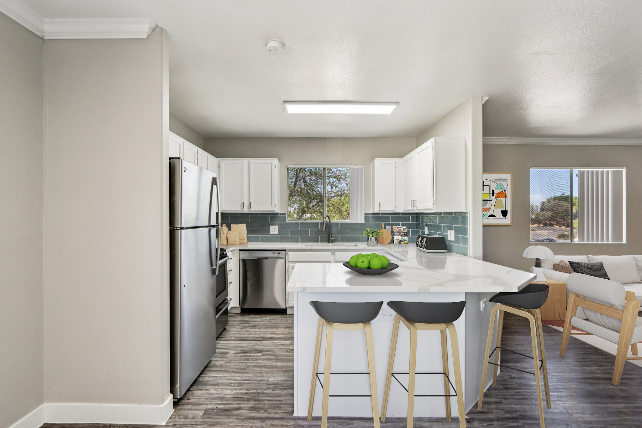 Modern kitchen featuring white cabinets, stainless steel appliances, and a blue tiled backsplash. A large island with bar stools is centrally located, and a bowl of green apples sits on the counter. The space includes a cozy seating area in the background with a light-colored sofa and a large window letting in natural light.
