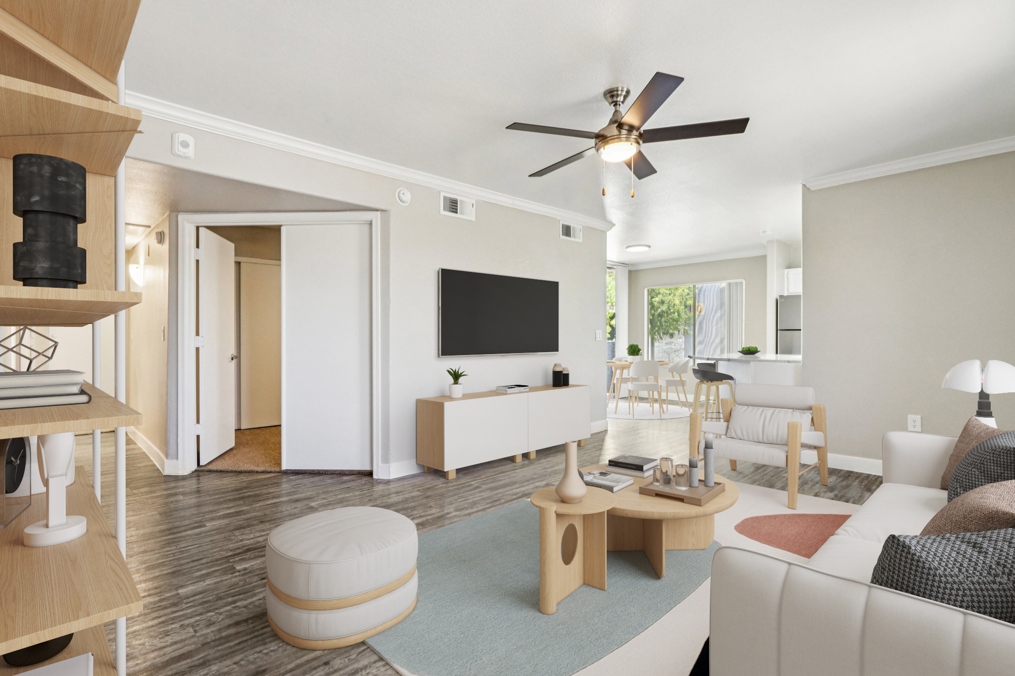 A modern, stylish living room with a neutral color palette. Features a comfortable white sofa, a wooden coffee table, and a light rug. A ceiling fan hangs above, and there is a large flat-screen TV mounted on the wall. Natural light streams in through a window, showcasing a dining area in the background.