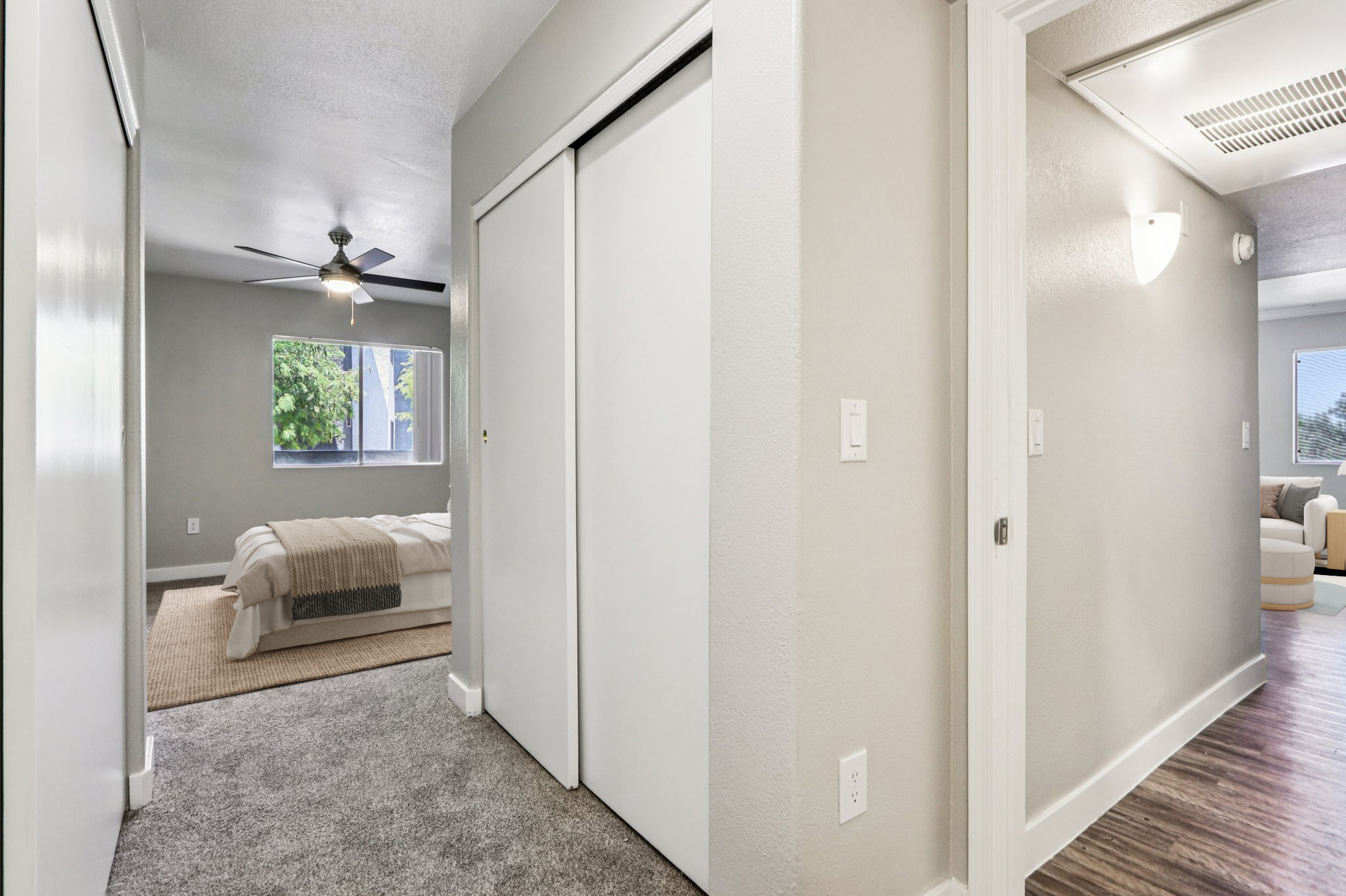 A hallway leading to a bedroom with a bed and window. The walls are painted gray, and there are closet doors on the left. A ceiling fan is visible, and light spills in from the window. The floor has carpet in the hallway and wood-like flooring in the main area.