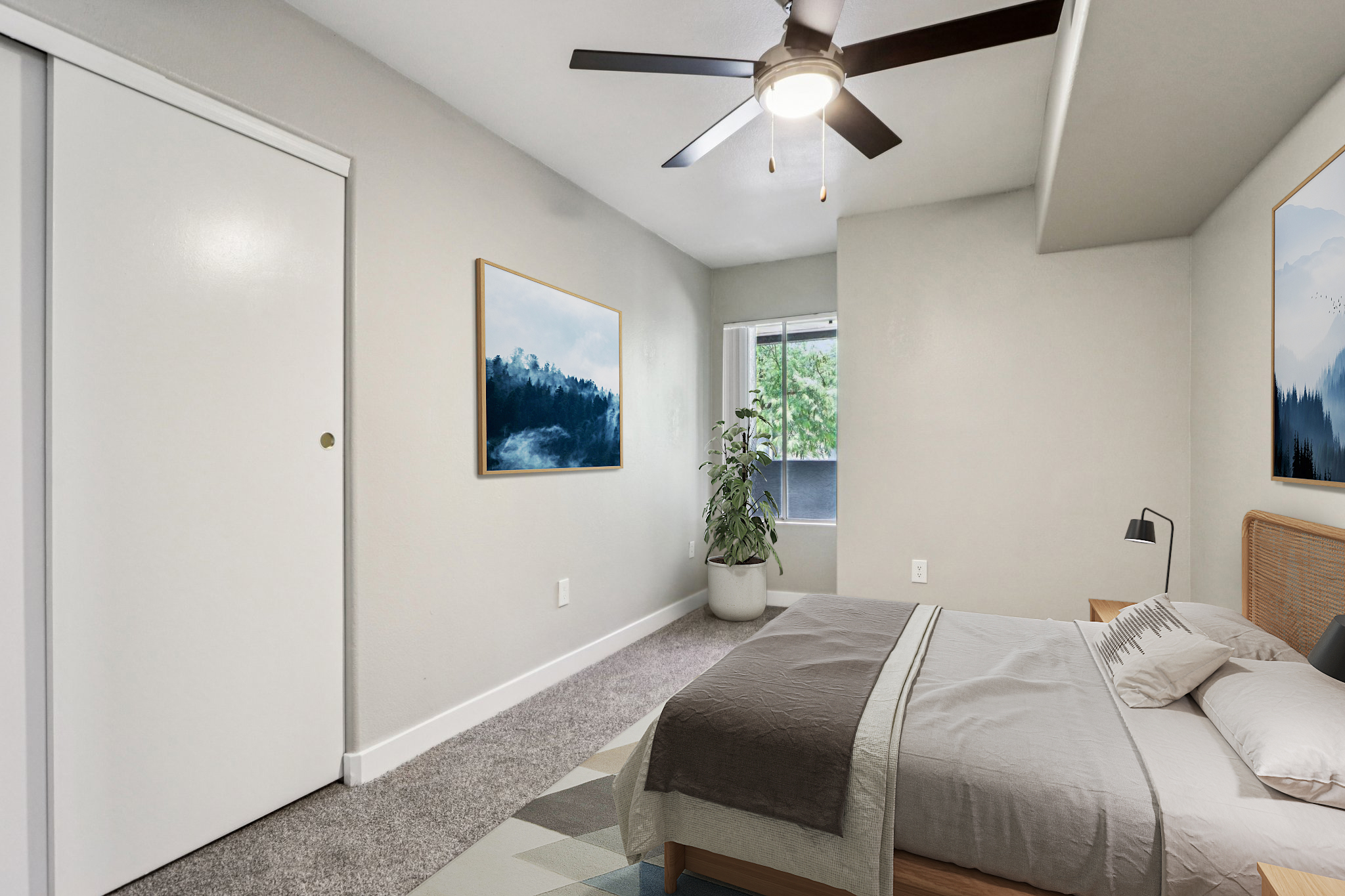 A cozy bedroom featuring a queen-sized bed with a light gray blanket, a ceiling fan, and two framed landscape paintings on the walls. A potted plant sits in the corner near a window, and there’s a white closet door visible. The room has neutral wall colors and a soft carpeted floor.