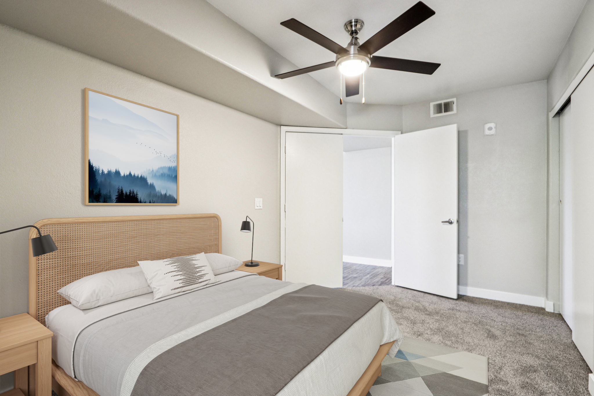 A cozy bedroom featuring a queen-sized bed with light gray bedding and a decorative throw pillow. There is a minimalist wooden nightstand with a lamp beside the bed. A piece of framed artwork depicting a misty landscape hangs on the wall. The room has neutral-toned walls, a ceiling fan, and a doorway leading to another space.