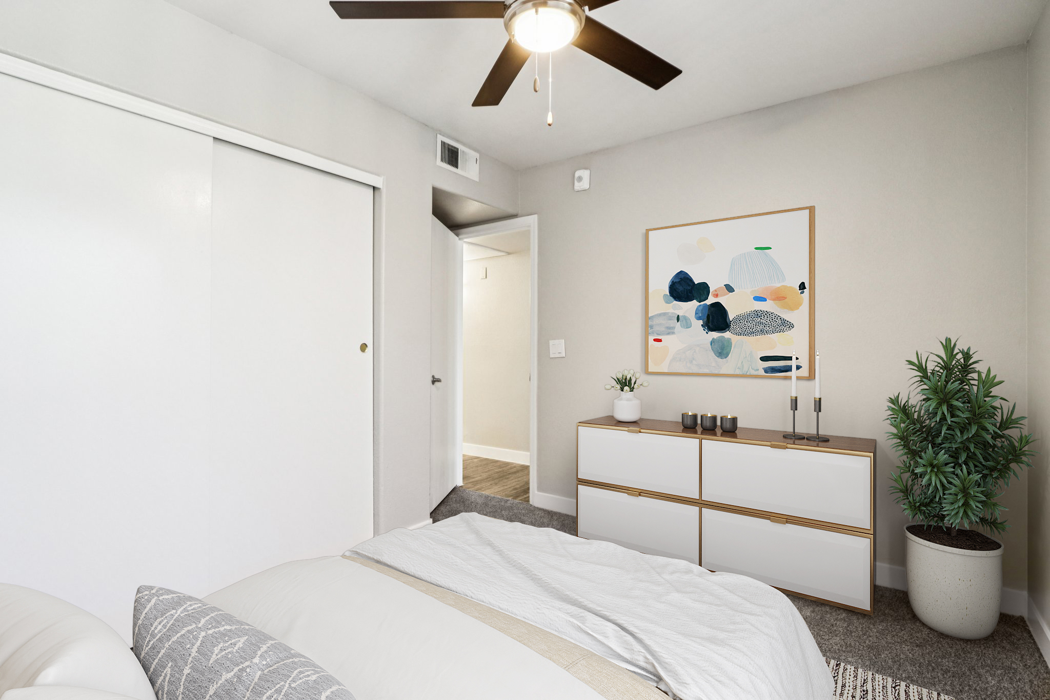 A modern bedroom featuring a large bed with a light-colored duvet, a white dresser with decorative items, and a potted plant. The walls are painted light gray, and there is a stylish ceiling fan. A piece of abstract art hangs above the dresser, complementing the contemporary decor. A door leads to another room in the background.