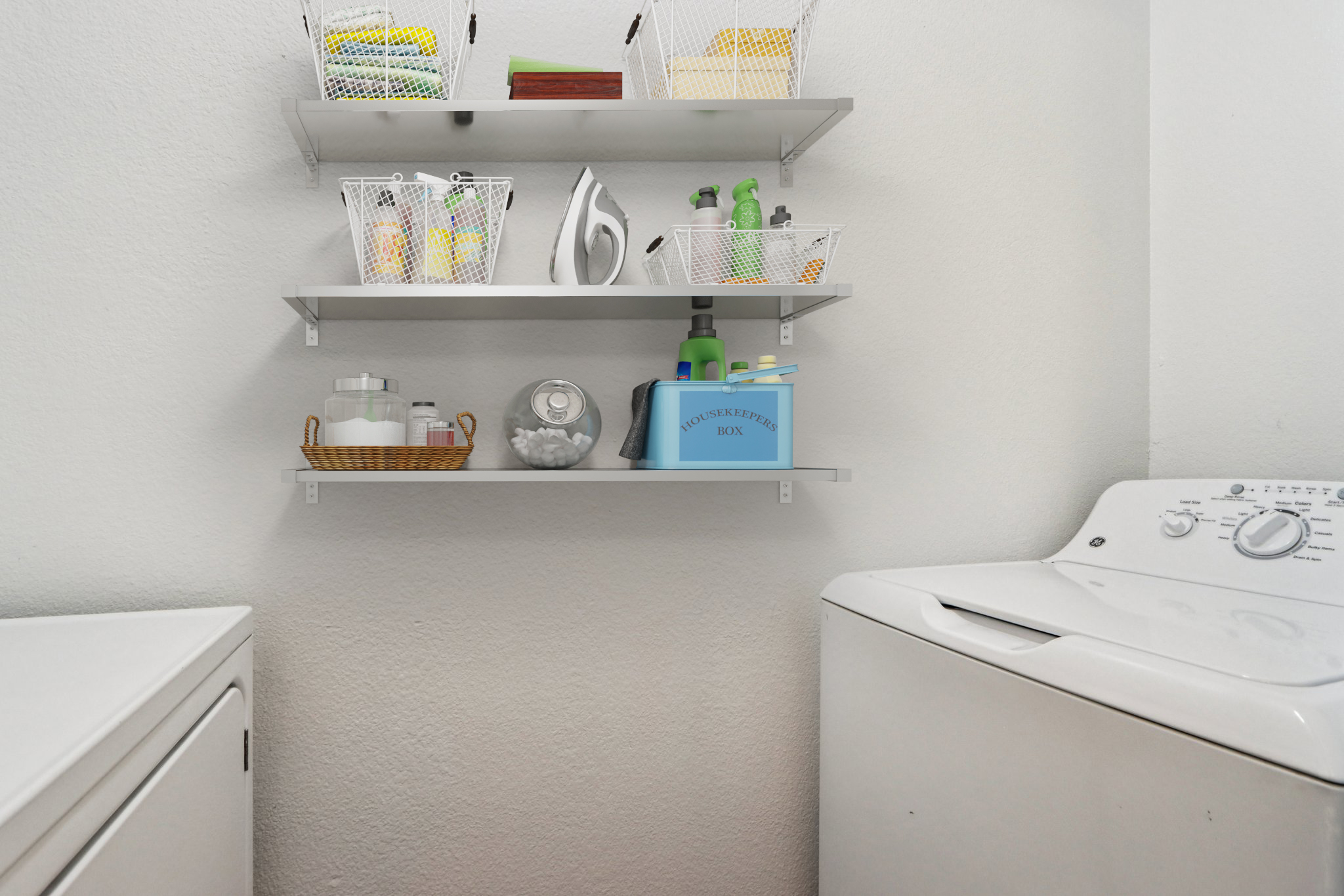 Shelves mounted on a wall in a laundry room, holding various cleaning supplies and storage baskets. A washing machine is visible on the left, and a blue box labeled "HOUSEHOLD BOX" is prominently placed on the shelf, along with a glass jar and a woven basket. The walls are plain and light-colored.