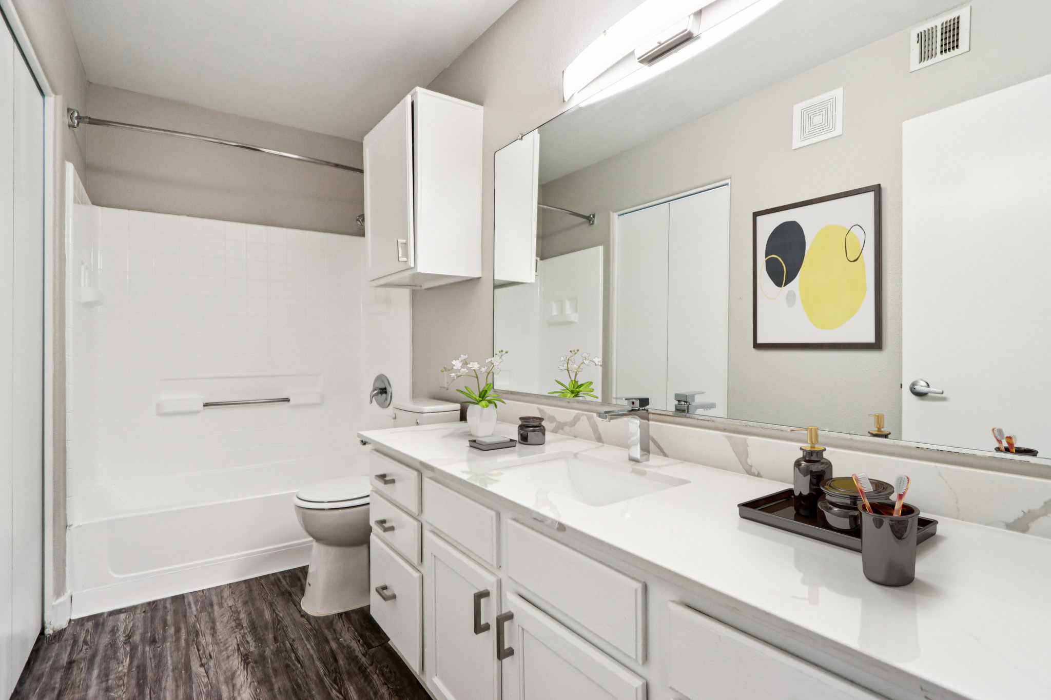 A modern bathroom featuring a white vanity with a sink, a large mirror above, potted plants, and decorative items on the counter. There is a white bathtub with a shower, light gray walls, and dark wood-like flooring, creating a clean and contemporary aesthetic.