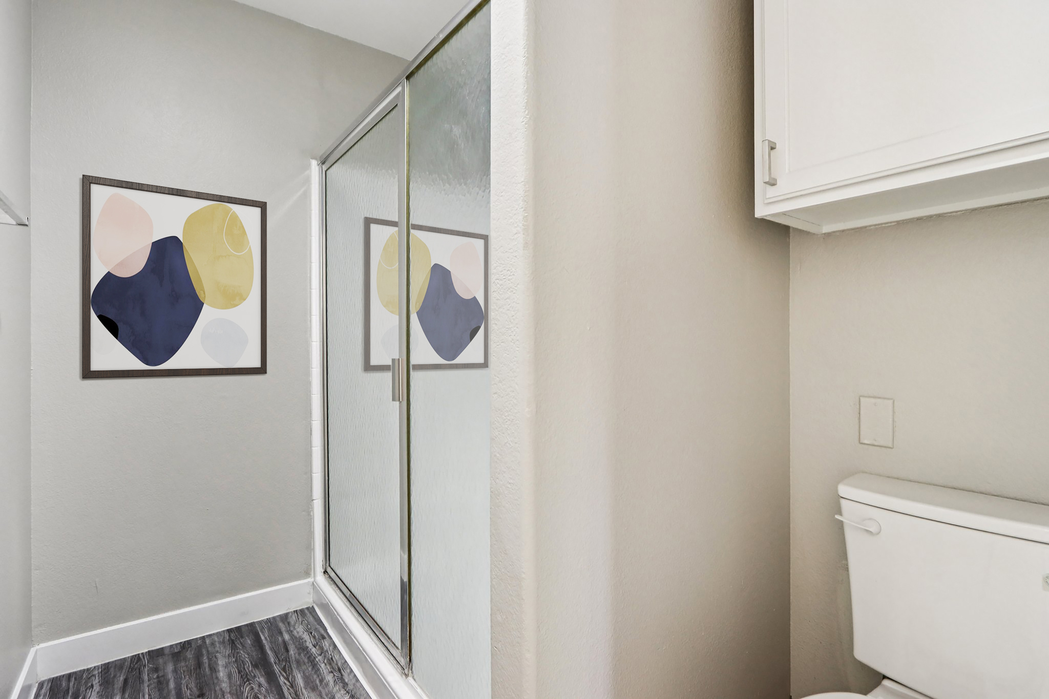 A modern bathroom featuring a glass shower stall, a toilet, and two abstract art pieces framed on the wall. The walls are painted in a light color, and the floor has dark laminate tiles. The space is well-lit and appears clean and contemporary.