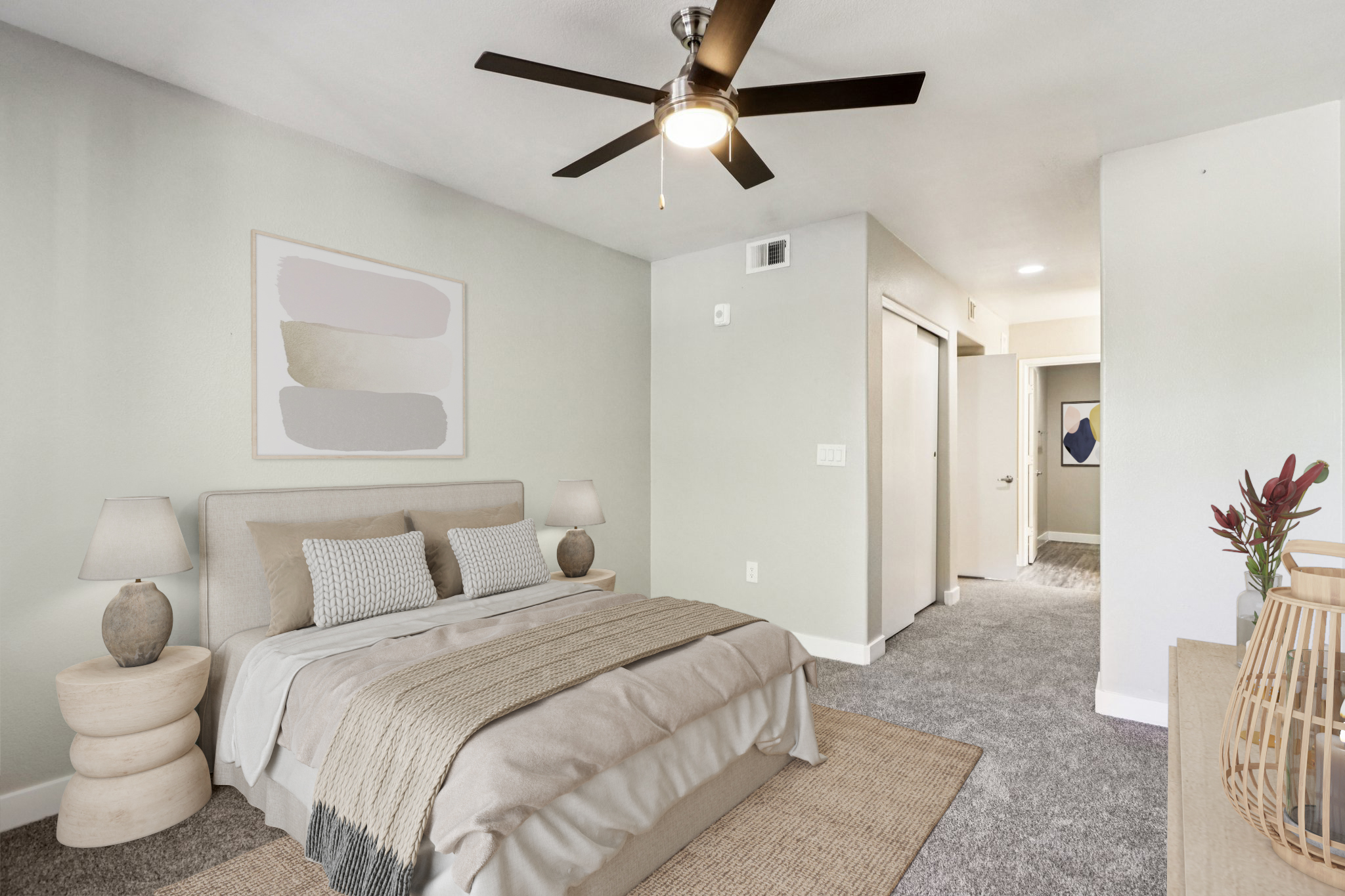 A modern bedroom featuring a light-colored wall with a ceiling fan. The room includes a bed with neutral bedding, two bedside lamps, and a cozy area rug. Adjacent to the bed is an open doorway leading to a hallway, with an abstract artwork visible on the wall. Decorative elements add a touch of warmth.