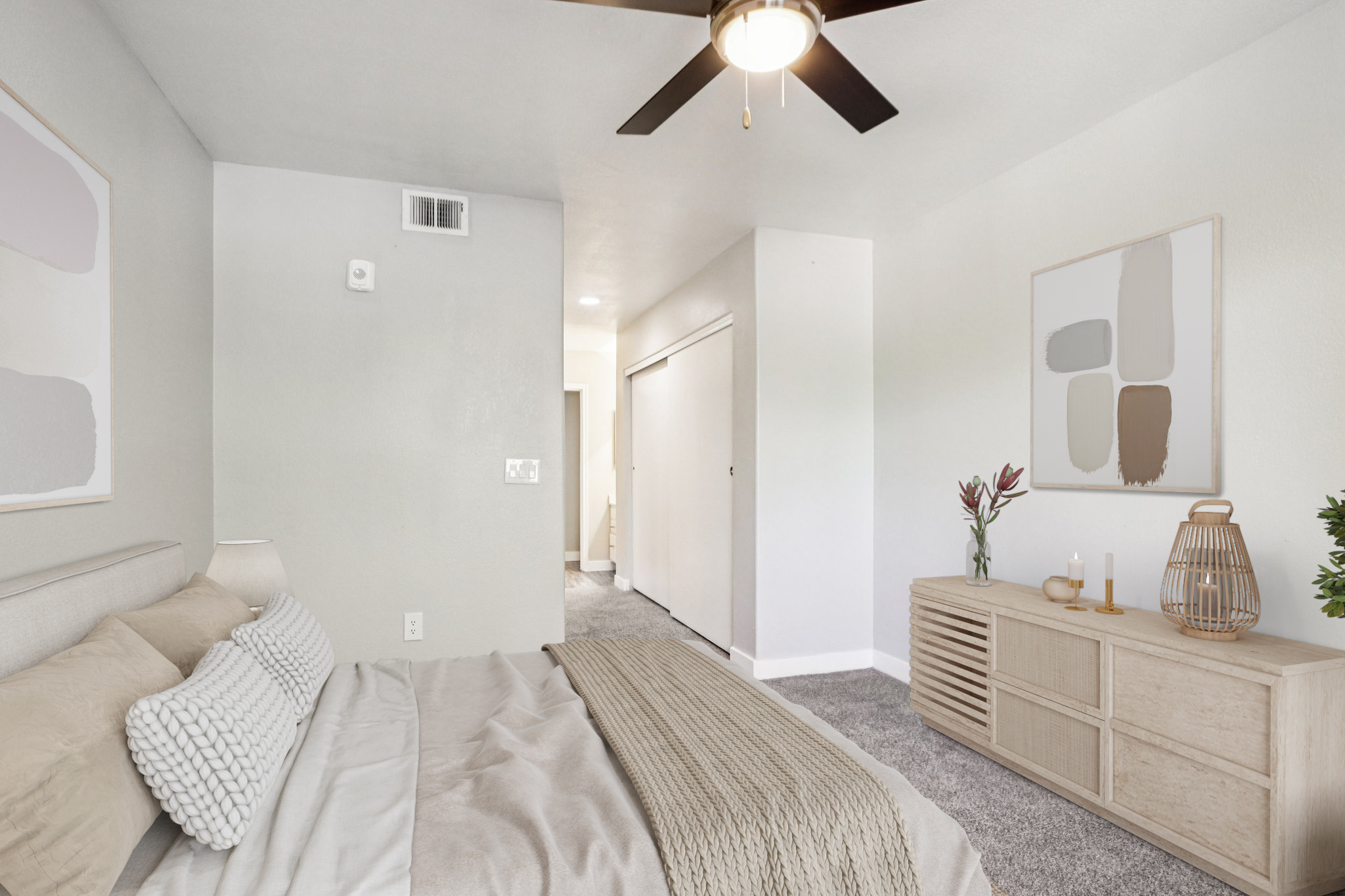 A cozy bedroom featuring a neutral color palette, a ceiling fan, and soft lighting. The room includes a bed with decorative pillows, a wooden sideboard, and a decorative plant. A doorway leads to an adjacent room, creating a spacious and inviting atmosphere.