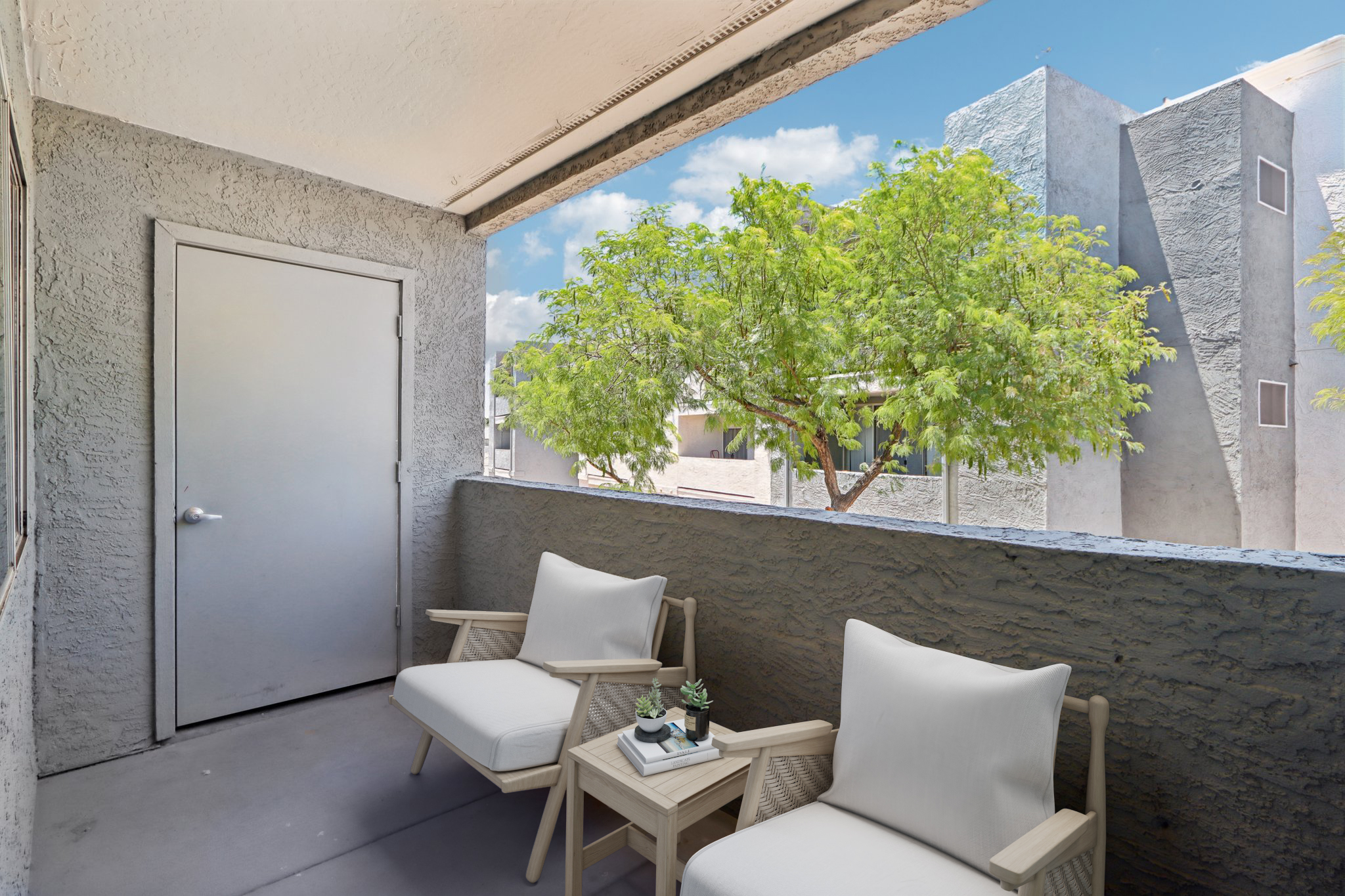 A balcony view featuring two white armchairs with a small side table. The space is adorned with a potted plant. In the background, there are trees and surrounding buildings under a bright blue sky with fluffy clouds.
