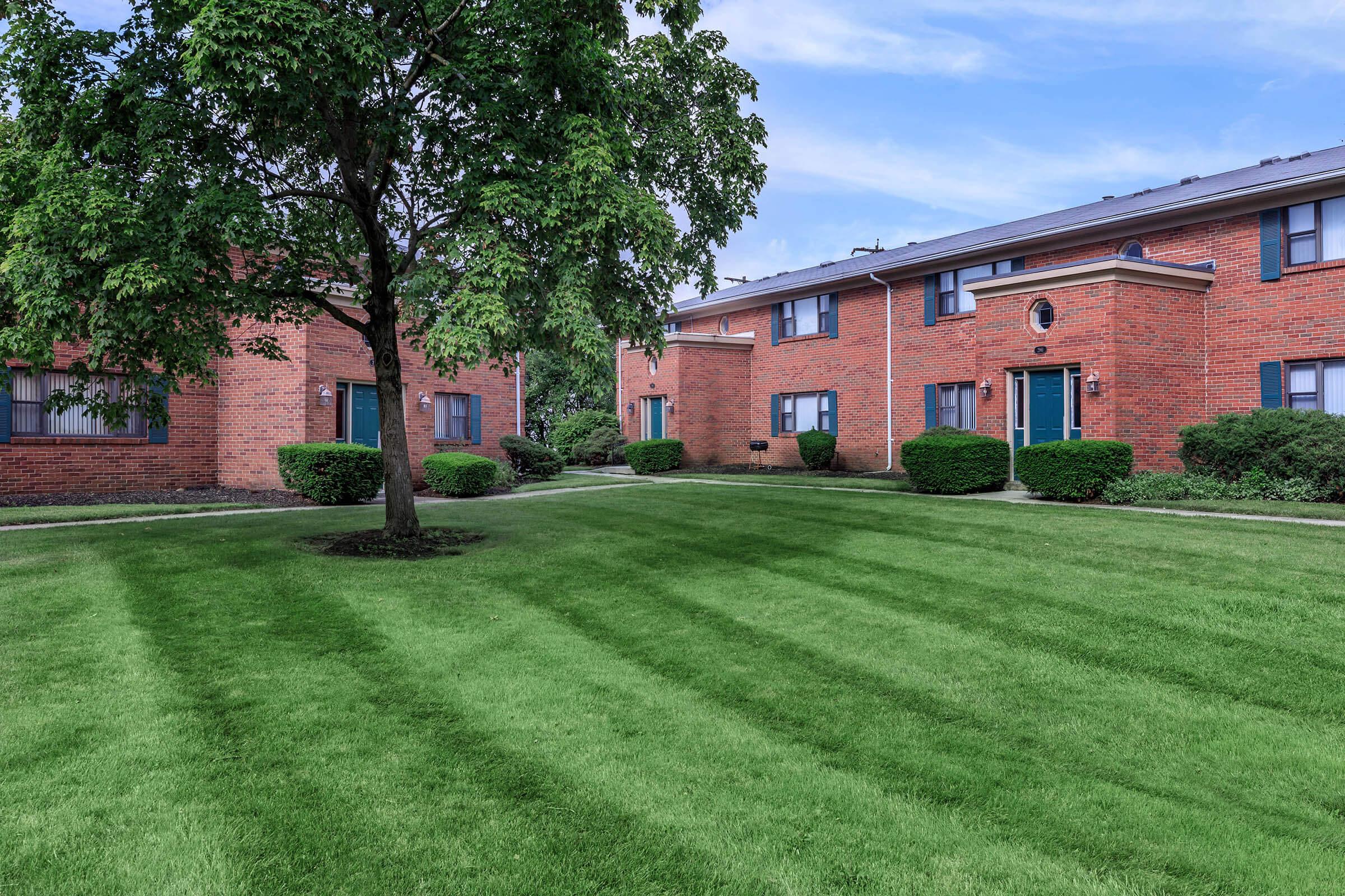 A well-maintained lawn with neatly mowed grass, surrounded by brick apartment buildings with green doors. Trees and shrubs enhance the landscape, creating a peaceful residential atmosphere on a clear day.