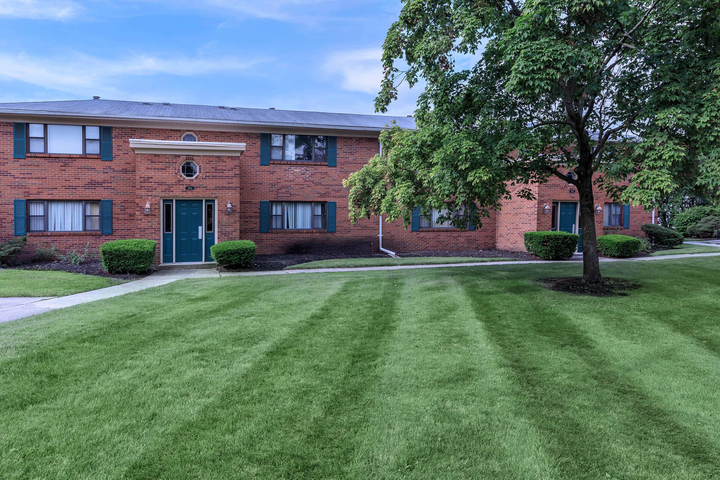 A well-maintained brick apartment building with green doors, surrounded by manicured lawns and neatly trimmed shrubs. The exterior features large windows and a tree providing shade in the foreground, with a clear blue sky in the background. Pathways lead to the entrance, enhancing the inviting atmosphere.
