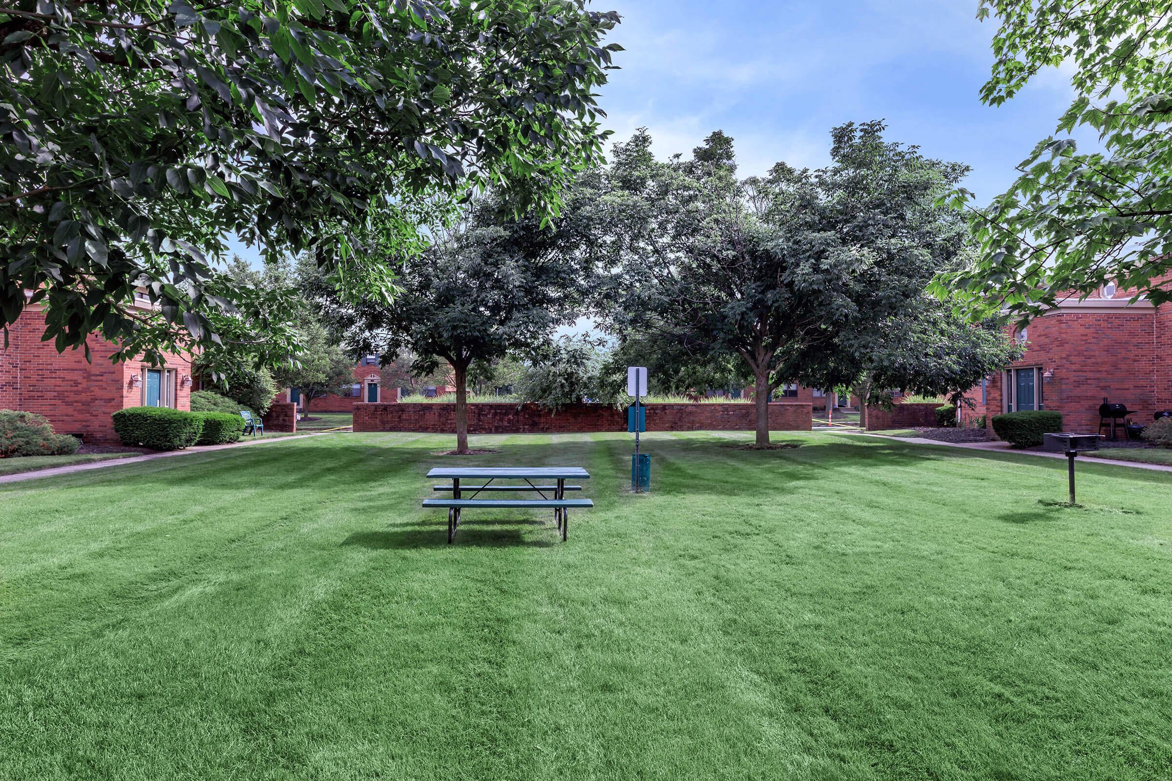 A well-maintained grassy area with a picnic table in the center, surrounded by green trees and red brick buildings. The scene is calm and inviting, featuring a clear blue sky and neatly trimmed landscaping.