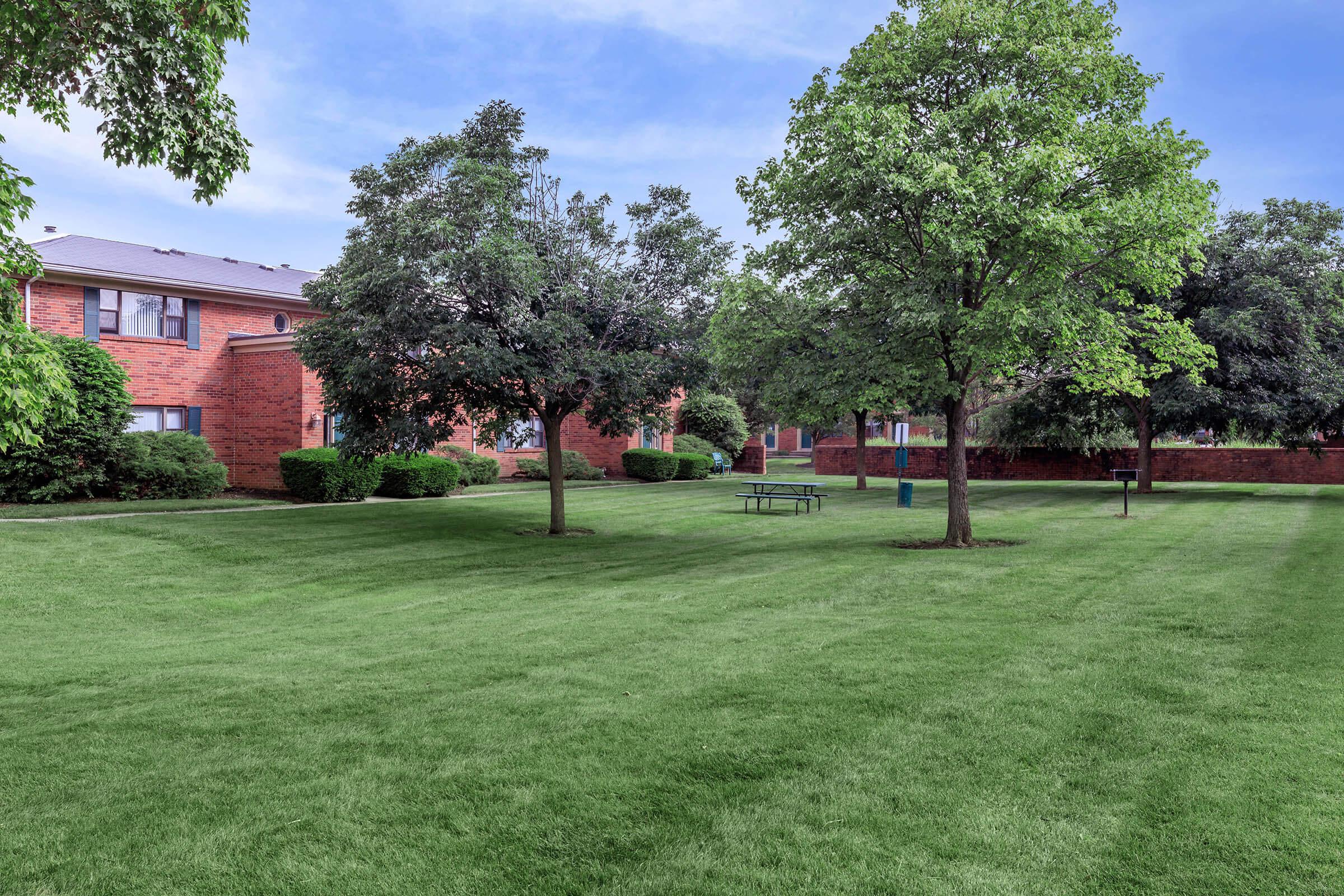 A well-maintained grassy area featuring trees, shrubs, and a picnic table. The surroundings include red brick buildings, providing a serene outdoor space for relaxation and recreation. The sky is clear, enhancing the peaceful atmosphere of the scene.