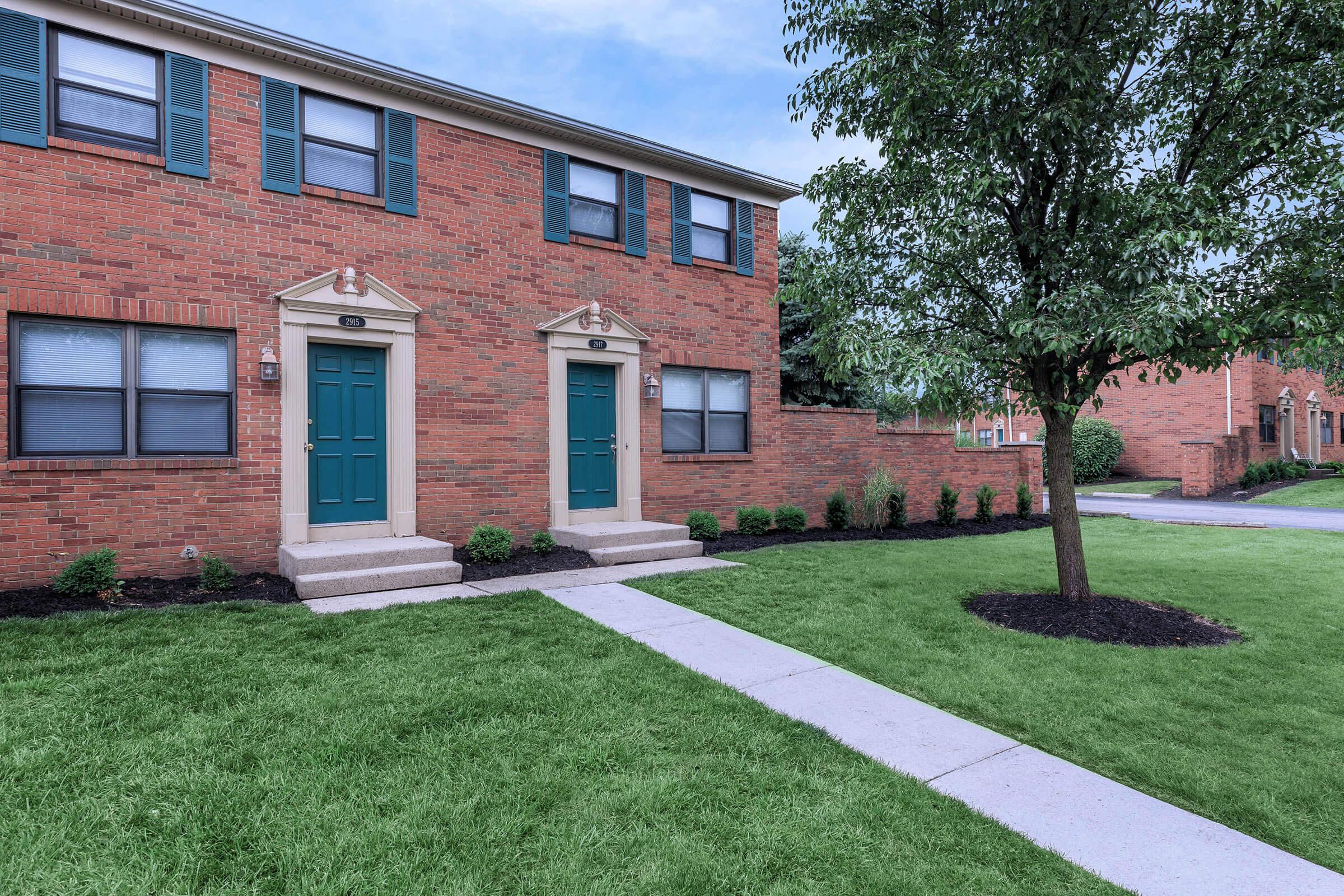 Two identical brick apartment entrances with green doors, flanked by small trees and manicured grass. A paved walkway leads to the doors, and additional apartment buildings can be seen in the background. The scene is well-kept and inviting, featuring a clear blue sky.