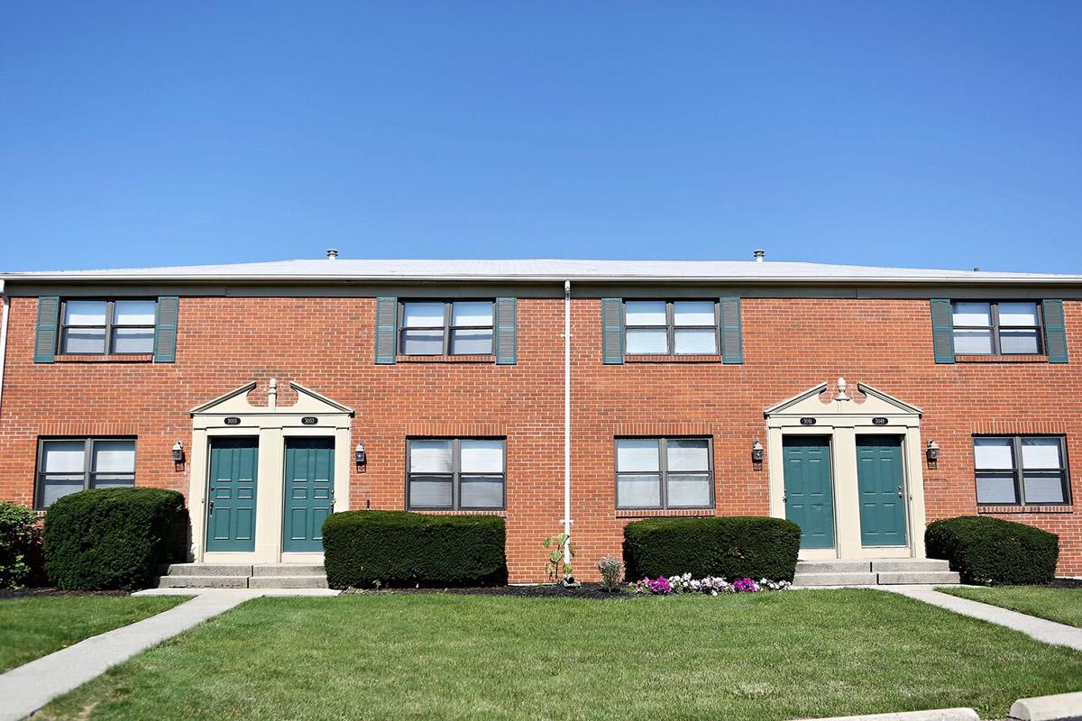 A brick apartment building featuring two identical units, each with green doors and shutters. The front yard is well-maintained with bushes and a flower bed. Clear blue sky above. Pathways lead to the entrances of the apartments.