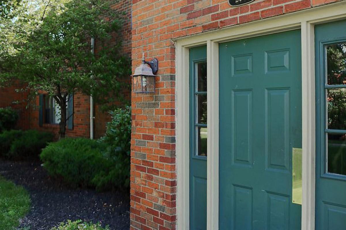 Exterior view of a brick building featuring a green double door with decorative lighting above. Surrounding foliage includes neatly trimmed shrubs and grass, contributing to a tidy landscape. The scene reflects a welcoming entrance.