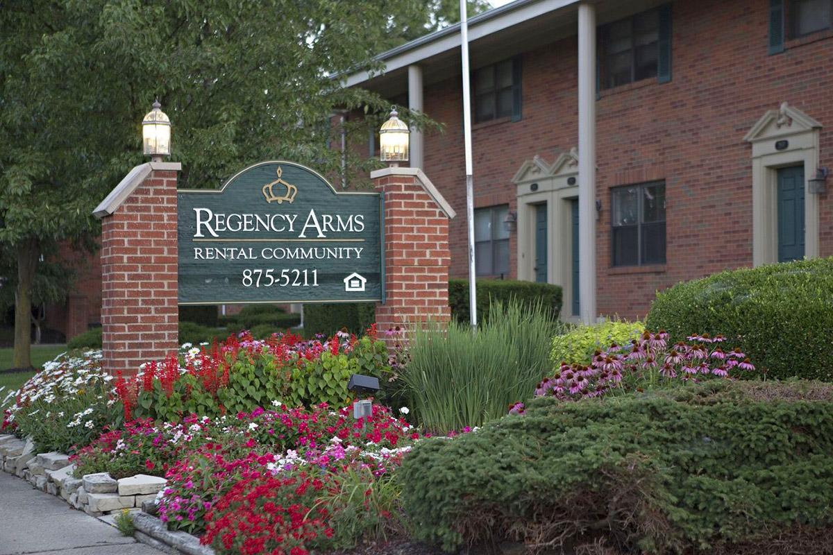 Sign for Regency Arms Rental Community surrounded by vibrant flower beds and landscaping, featuring a brick structure with lamps. The sign includes the phone number 875-5211.