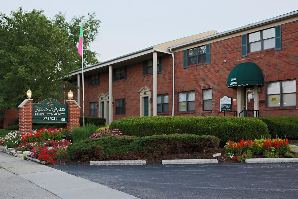 Exterior view of Regency Arms Rental Community, featuring a brick building with a green awning over the office entrance. Colorful flower beds line the entrance, and a sign indicates the property name and contact information. Lush greenery surrounds the area, creating a welcoming environment.