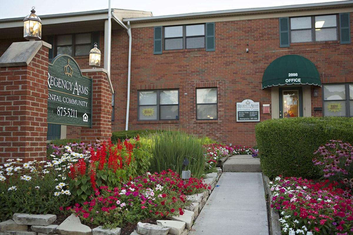 Sign for Regency Arms Rental Community in front of a brick building adorned with colorful flower beds. Visible are flower arrangements with red, pink, and white blooms, along with a paved walkway leading to the entrance, which features a green awning and signage indicating the office location.