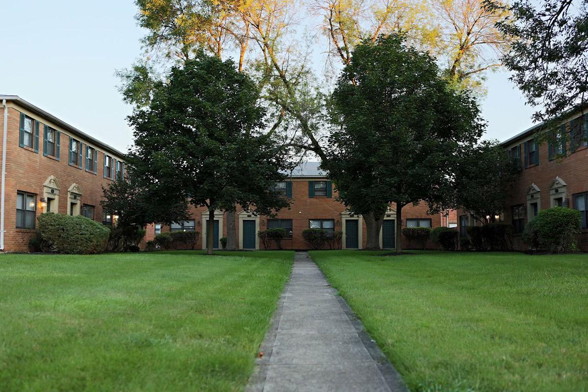 A peaceful residential area featuring a well-maintained courtyard surrounded by brick apartment buildings. Green lawns and trees frame a central path leading through the space, creating a serene atmosphere in the early morning light.