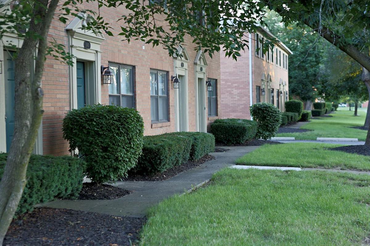 Row of residential buildings with brick facades and neatly trimmed bushes along a concrete walkway. Well-maintained grassy areas flank the path, creating a tidy and inviting atmosphere.