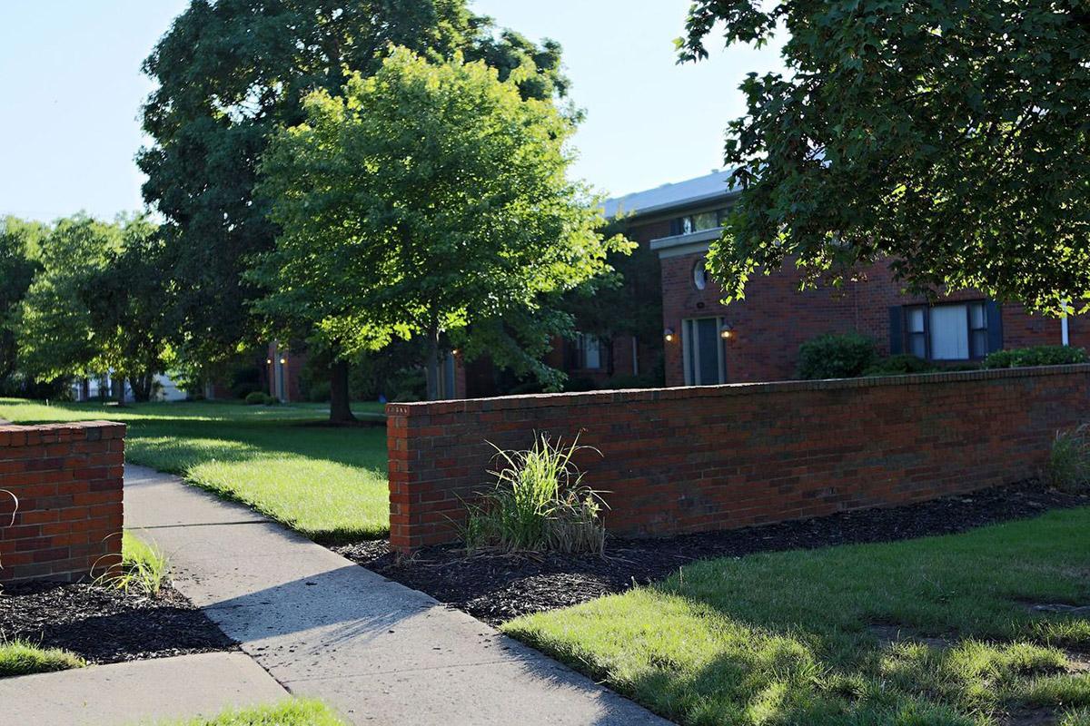 A peaceful residential scene featuring well-maintained brick apartments set amidst green lawns and trees. A pathway curves through the area, bordered by low brick walls and patches of mulch, inviting a serene atmosphere in a suburban neighborhood.
