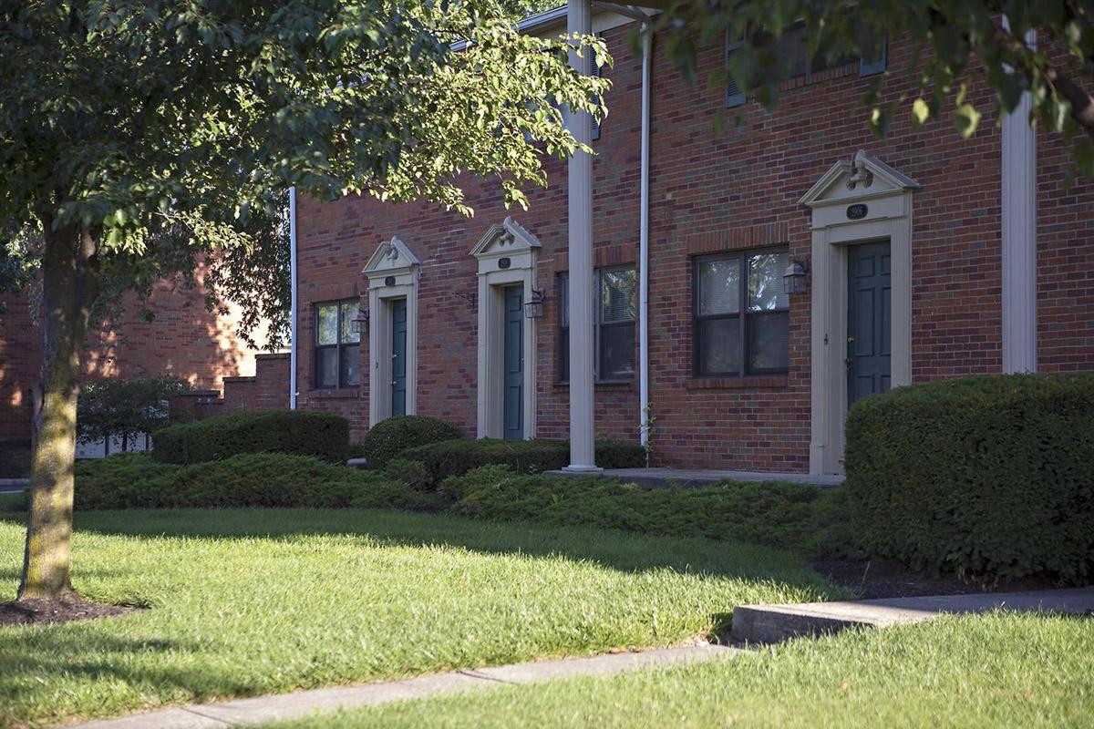 A quiet residential area featuring brick apartment buildings with green front doors, surrounded by neatly trimmed hedges and a well-maintained lawn. Sunlight filters through the trees, casting gentle shadows on the walkway.