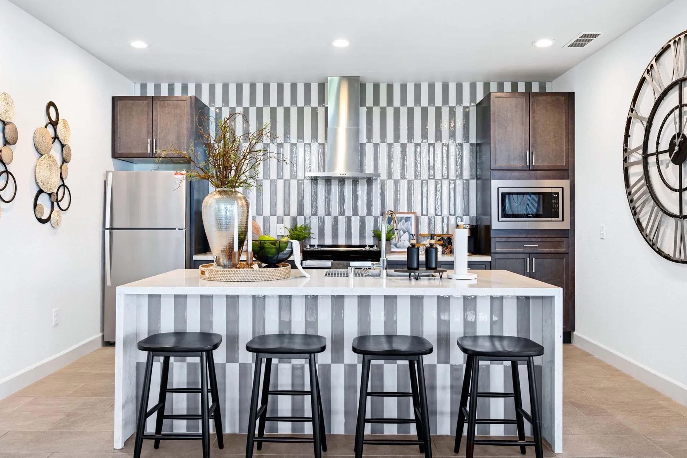 Modern kitchen featuring a large white island with four black stools, stainless steel appliances, and dark wood cabinetry. The walls are decorated with striped tiles and decorative wall art. A sleek metal clock is visible on the wall, adding to the contemporary aesthetic. Bright lighting enhances the inviting atmosphere.